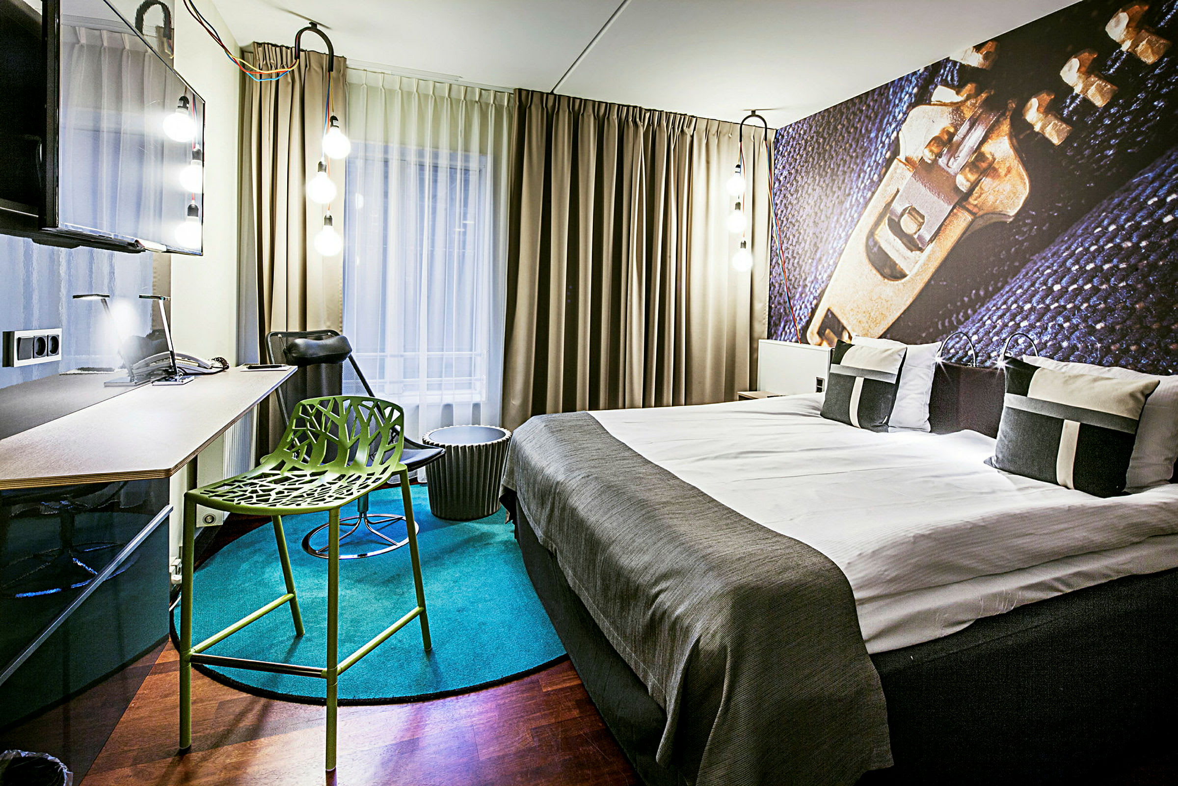 Modernisti sisustettu hotellihuone, jossa on parivuode, kirjoituspöytä, vihreä tuoli ja suuret verhot ikkunan edessä. (Tekoälyn luoma teksti)