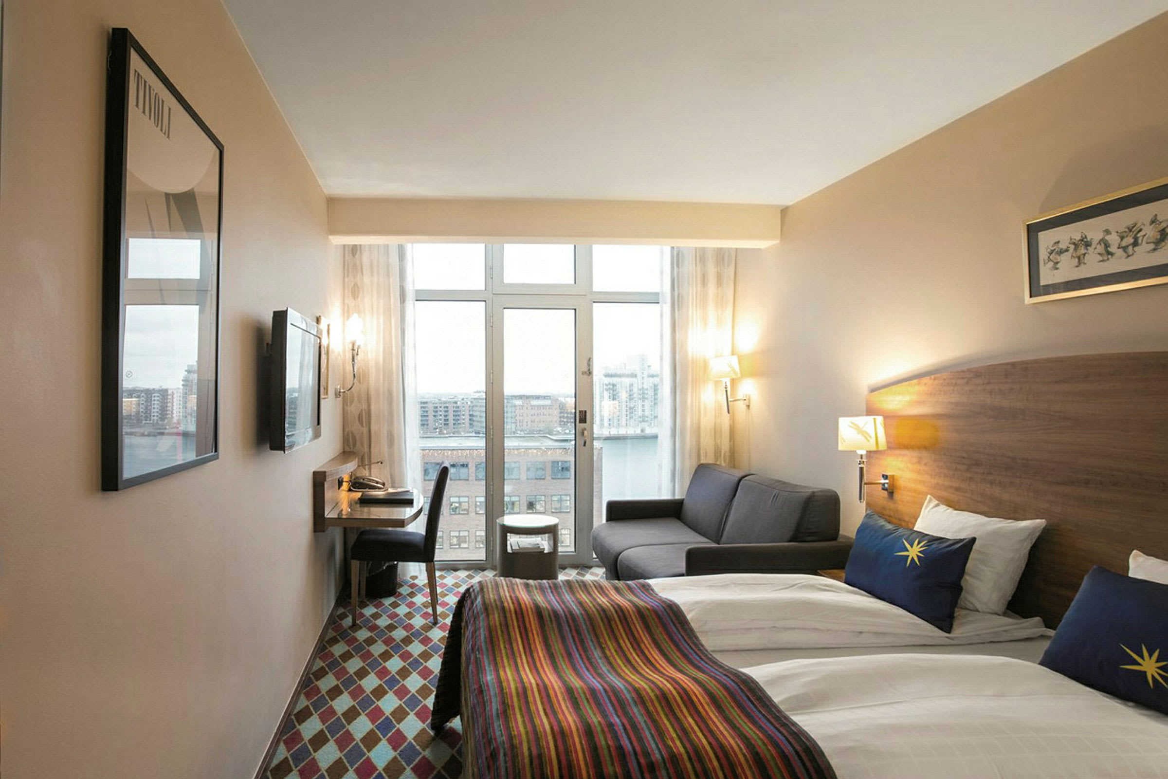 Hotellihuone, jossa on kaksi erillistä vuodetta, valkoiset lakanat ja värikkäät päiväpeitteet. Huoneessa on myös harmaa sohva, kirjoituspöytä tuoleineen ja näkymä kaupunkiin suurista ikkunoista. (Tekoälyn luoma teksti)