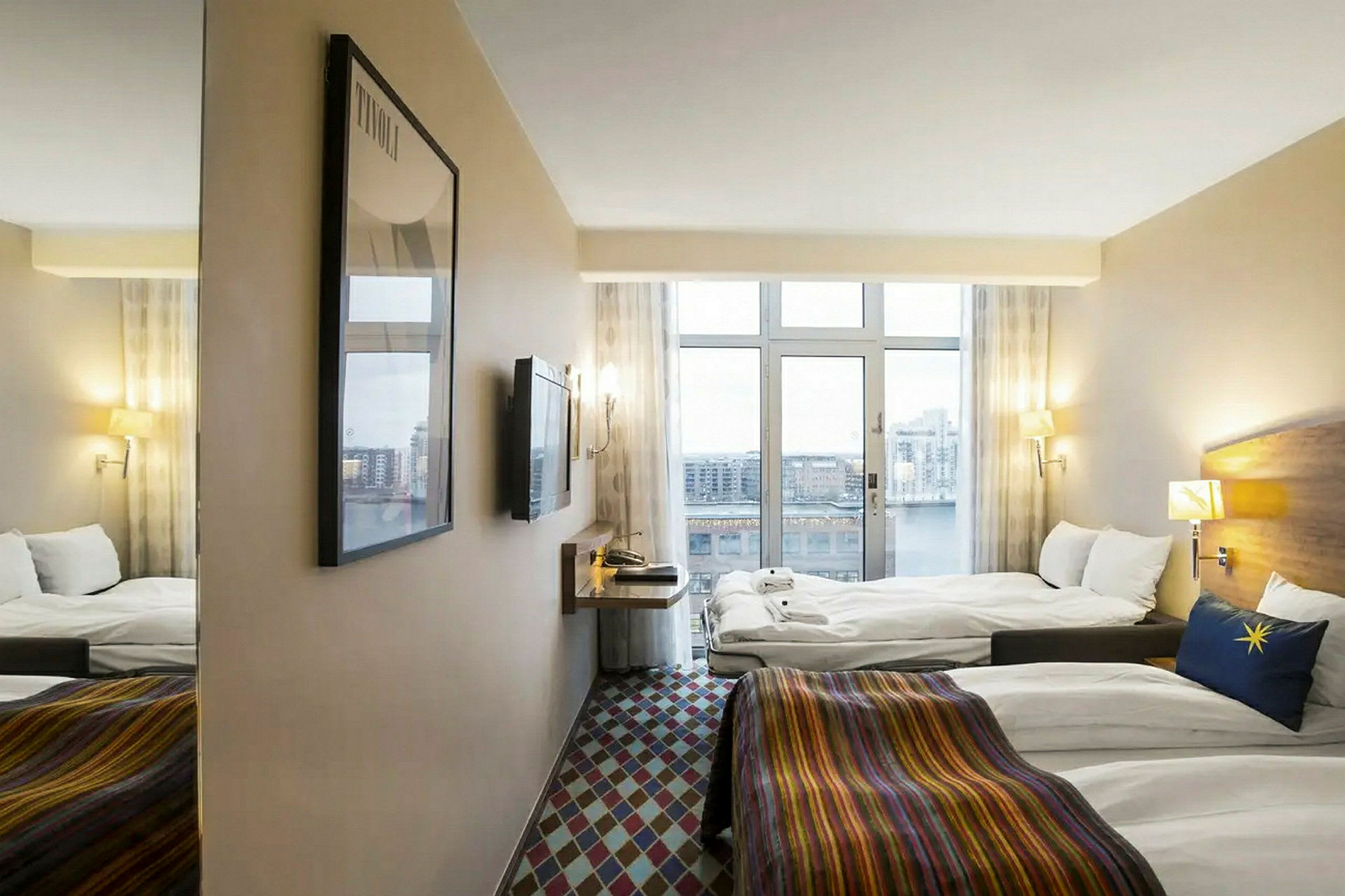 Hotellihuone, jossa on ikkunat, jotka tarjoavat näkymän kaupunkimaisemaan. Huoneessa on kaksi sänkyä, seinälle kiinnitetty TV ja peili. (Tekoälyn luoma teksti)
