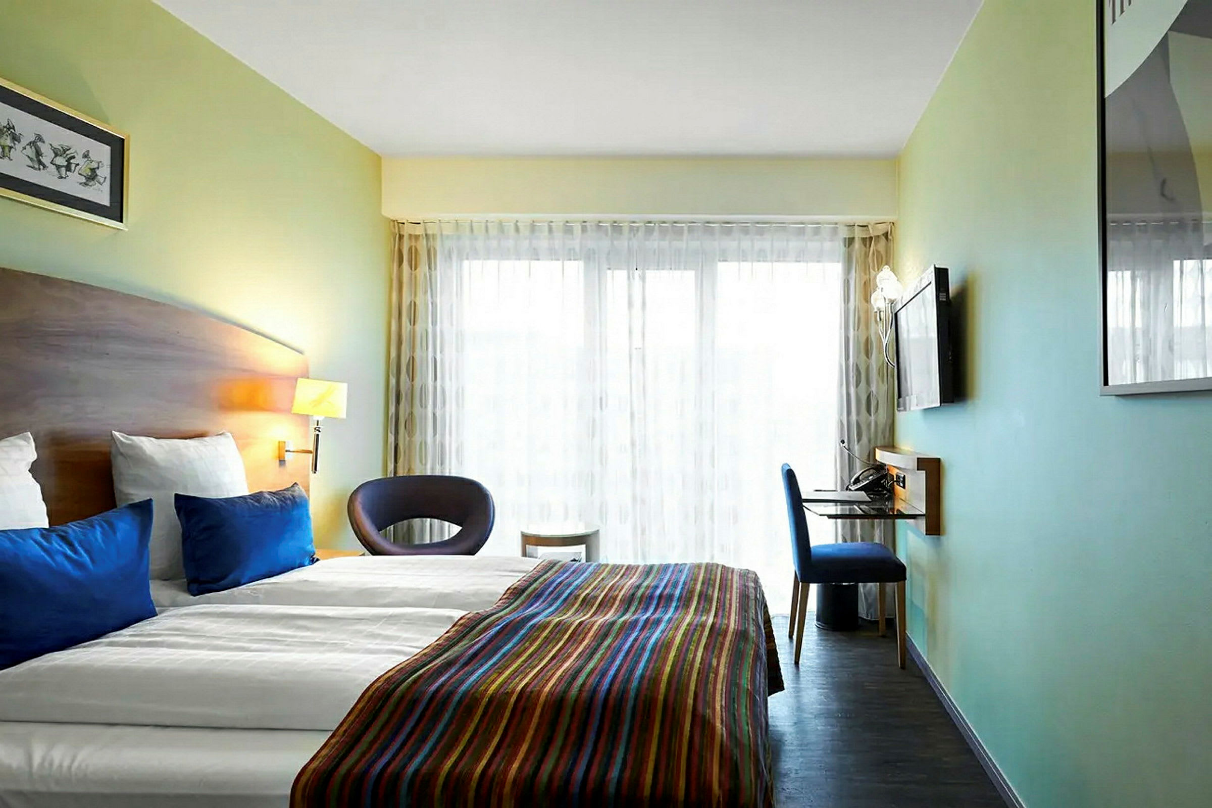 Nykyaikainen hotellihuone, jossa on parivuode, siniset tyynyt ja värikäs päiväpeite. Huoneessa on myös seinään kiinnitetty televisio, kirjoituspöytä ja tuoli sekä suuret ikkunat, joissa on vaaleat verhot. (Tekoälyn luoma teksti)