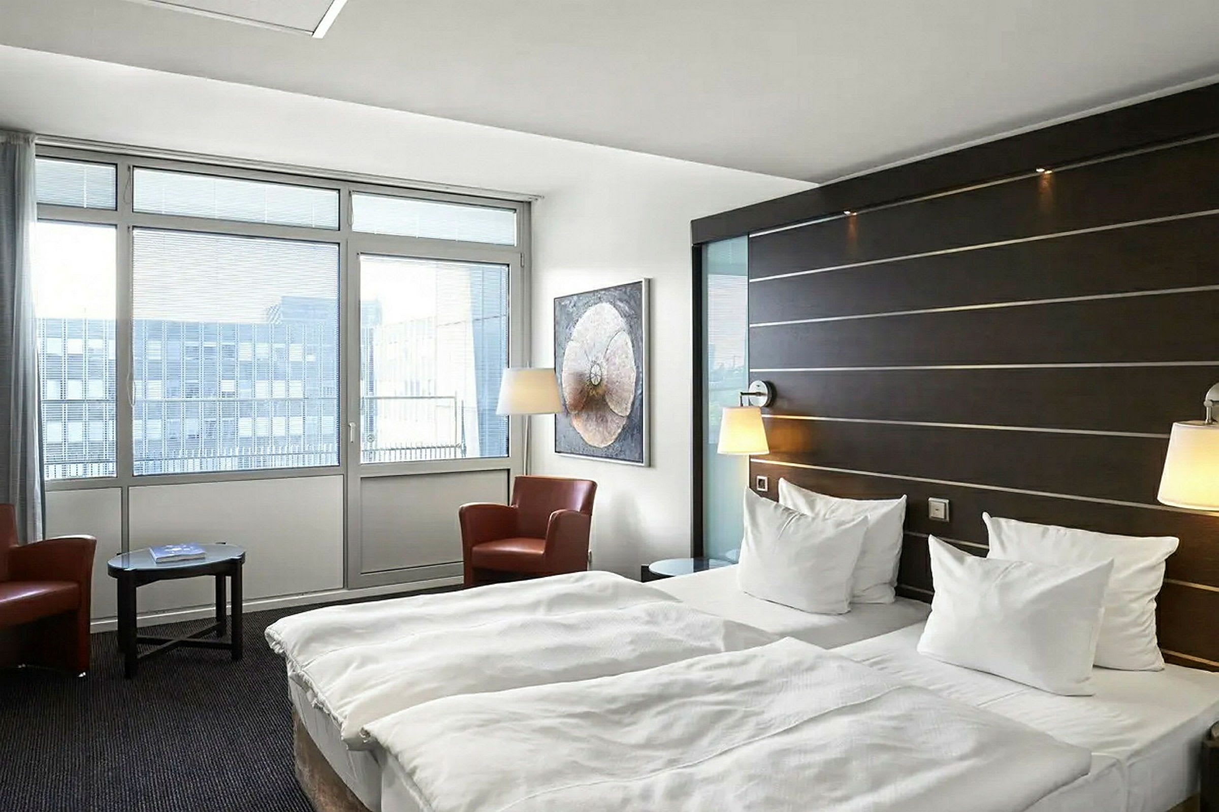Hotellihuone, jossa on kaksi erillistä vuodetta, istumaryhmä kahdella nojatuolilla ja pöytä sekä suuret ikkunat, joista tulvii luonnonvaloa. (Tekoälyn luoma teksti)