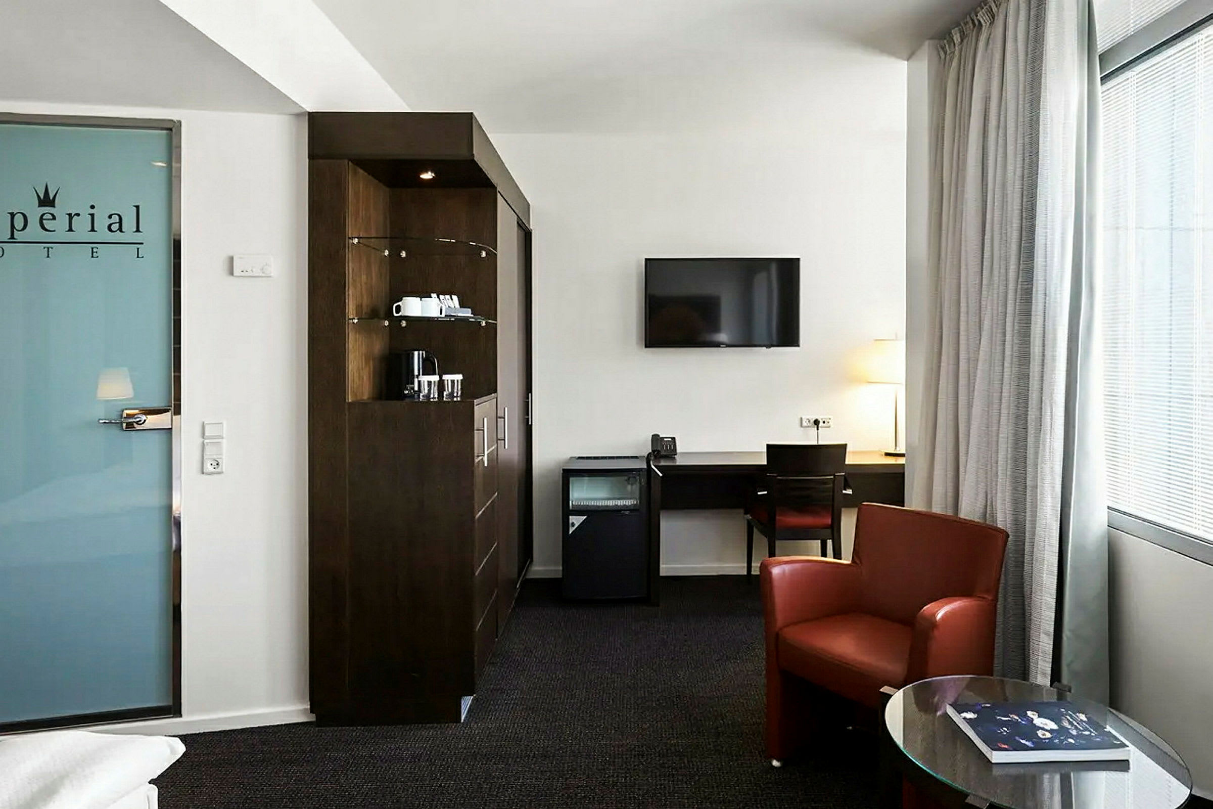 Moderni hotellihuone, jossa on huonekaluja, kuten työpöytä, punainen nojatuoli ja televisio. Huoneessa on myös minibaari ja kahvinkeitin. (Tekoälyn luoma teksti)