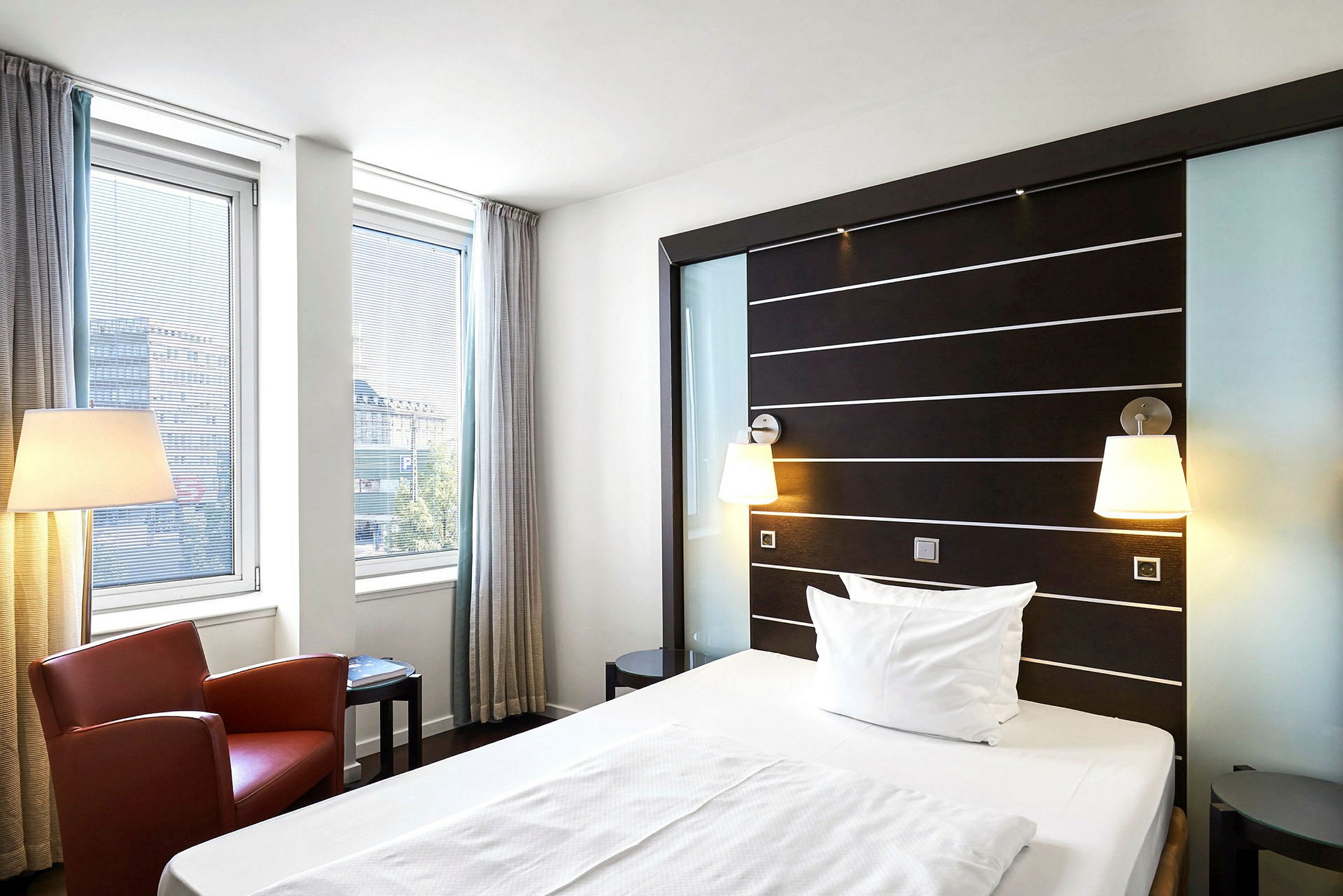 Moderni hotellihuone, jossa on yhden hengen vuode, kaksi ikkunaa verhoilla, punainen nojatuoli ja lattiavalaisin. (Tekoälyn luoma teksti)