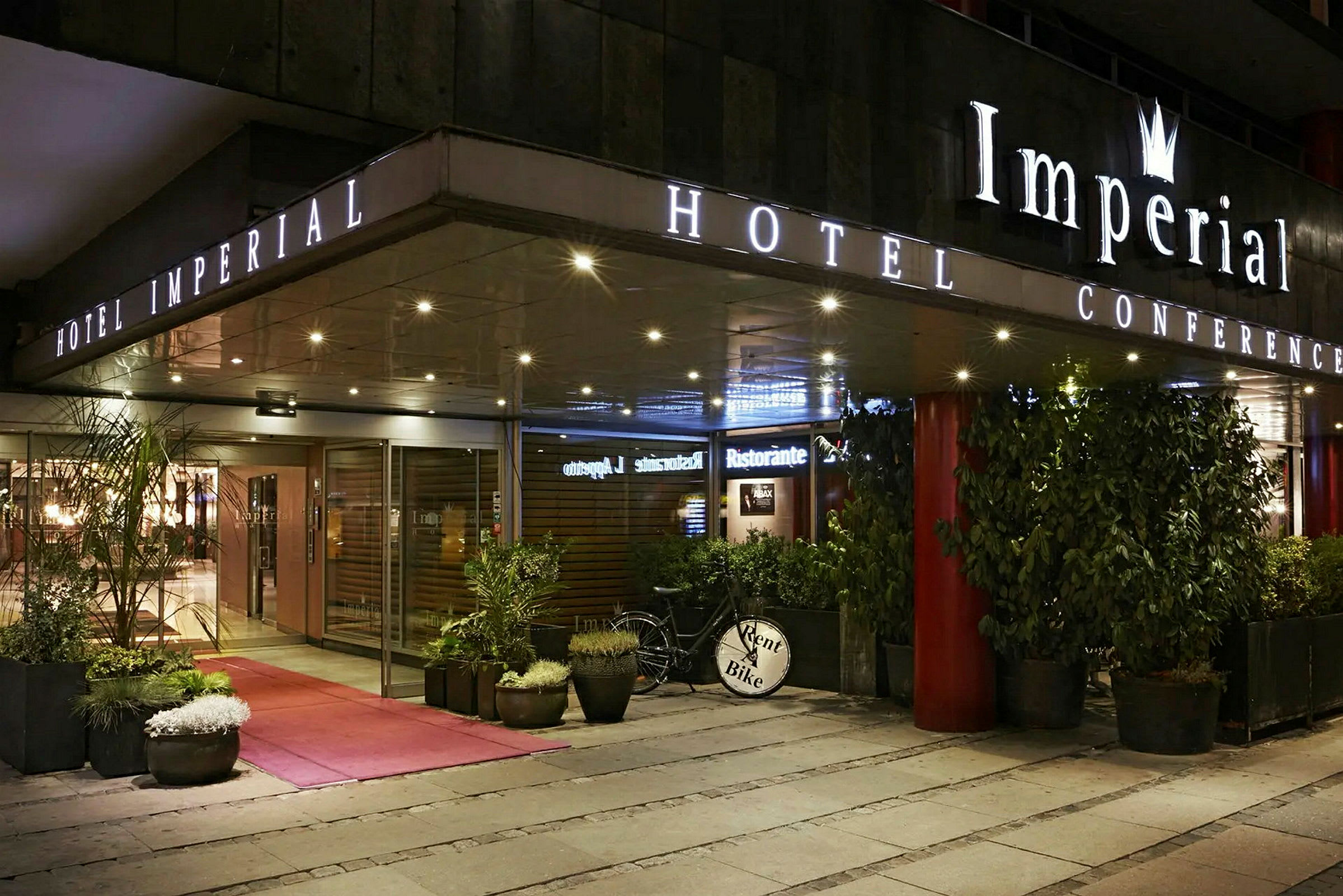 Hotel Imperial Conference -hotellin sisäänkäynti lasiovilla, ruukkukasveilla ja polkupyörällä, jossa on kyltti 'Rent a Bike'. (Tekoälyn luoma teksti)