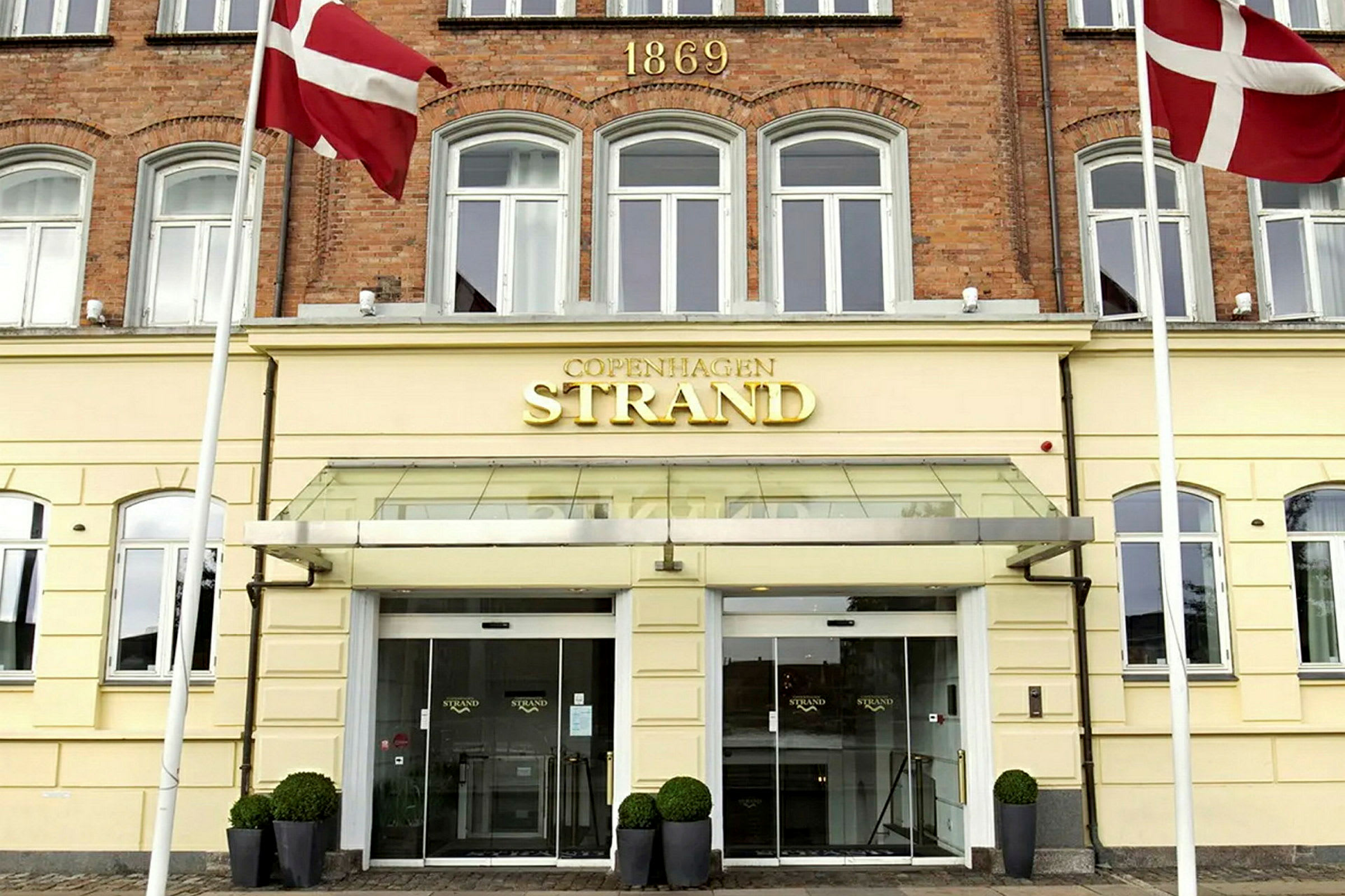 Rakennus, jossa on keltainen julkisivu ja kyltti, jossa lukee "Copenhagen Strand" sisäänkäynnin yläpuolella. Kaksi Tanskan lippua roikkuu sivuilla. (Tekoälyn luoma teksti)