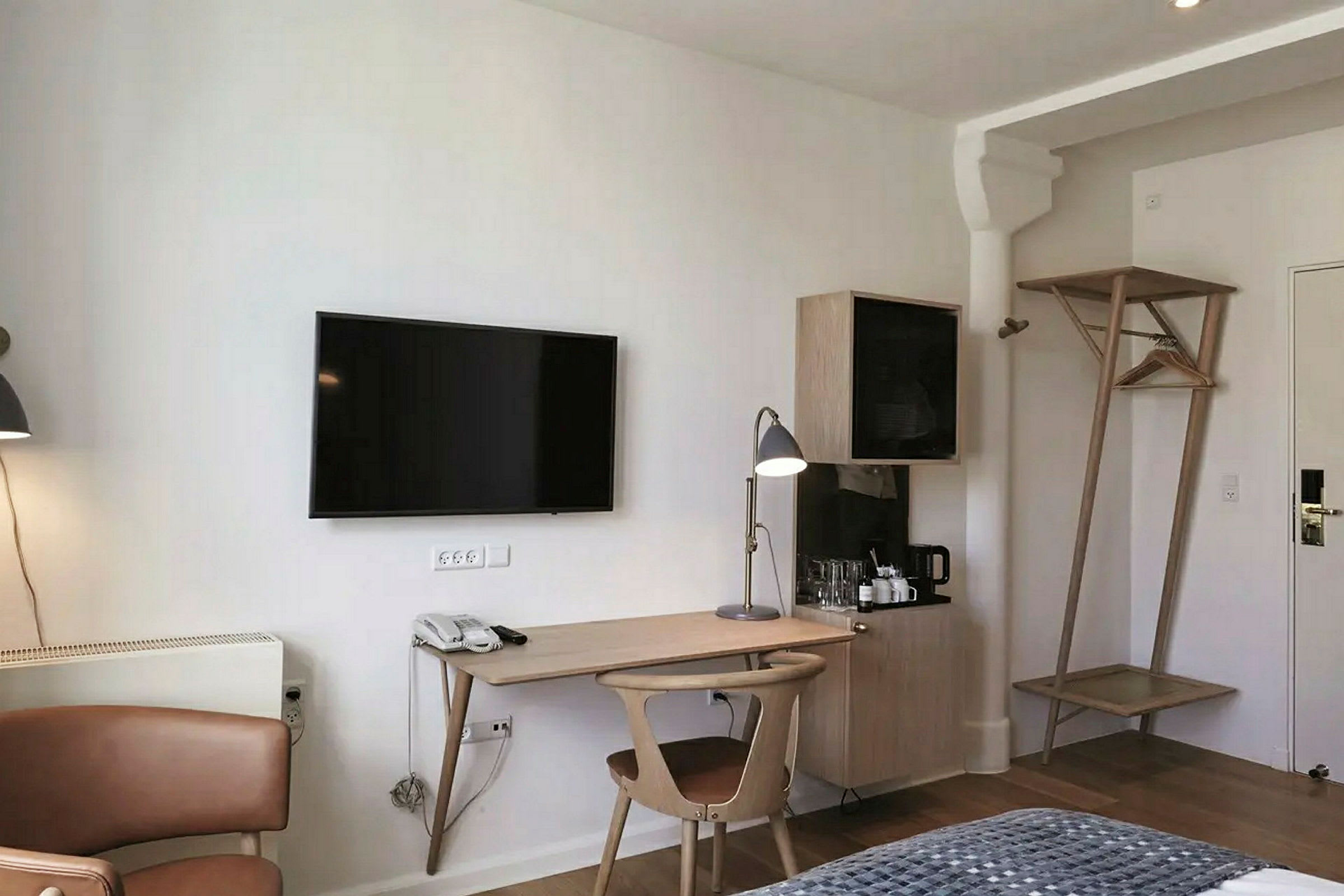 Hotellihuone, jossa on työtilaa, sisältäen kirjoituspöydän, tuolin ja pöytälampun. TV on kiinnitetty seinään yläpuolella. (Tekoälyn luoma teksti)