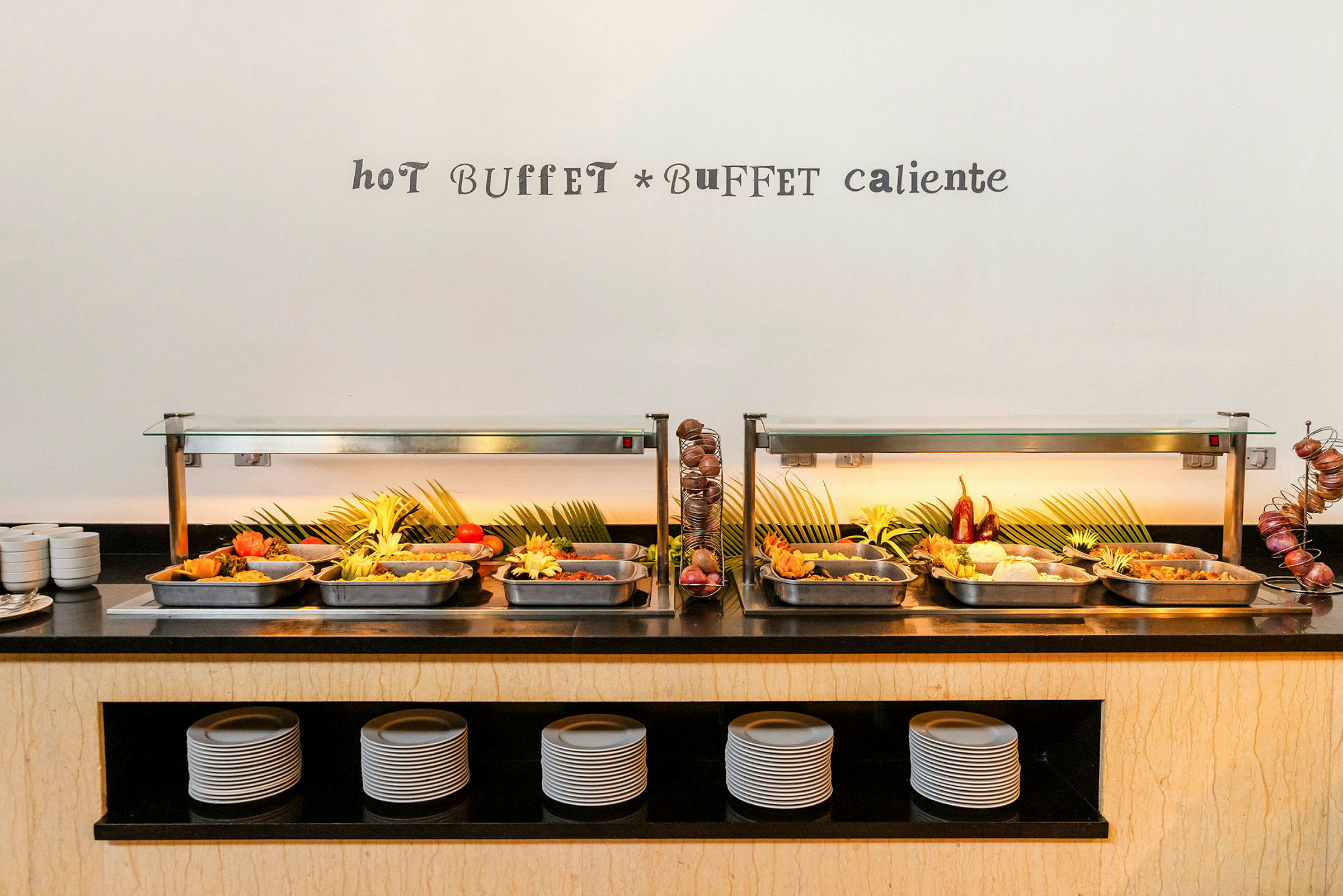 Lämmin buffet, jossa on erilaisia ruokalajeja tarjoiltuna teräsastioissa, koristeltu vihreillä lehdillä ja tuoreilla kasviksilla. (Tekoälyn luoma teksti)