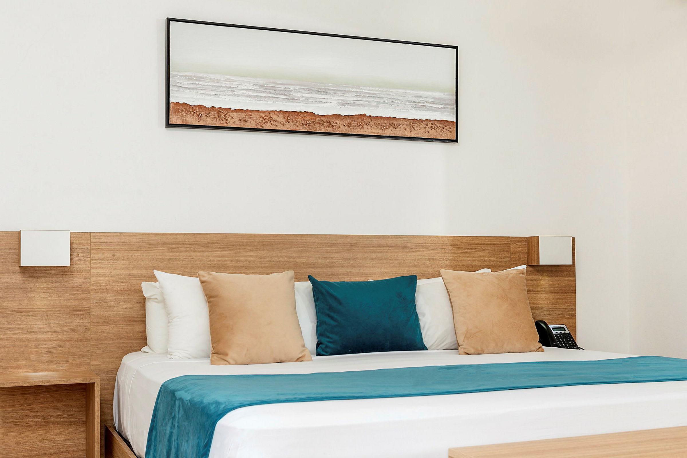 Hotellihuone, jossa sänky, joka on varustettu beigellä ja sinisillä tyynyillä sekä sinisellä päiväpeitteellä. Seinällä sängyn yläpuolella on maisemataulu. (Tekoälyn luoma teksti)