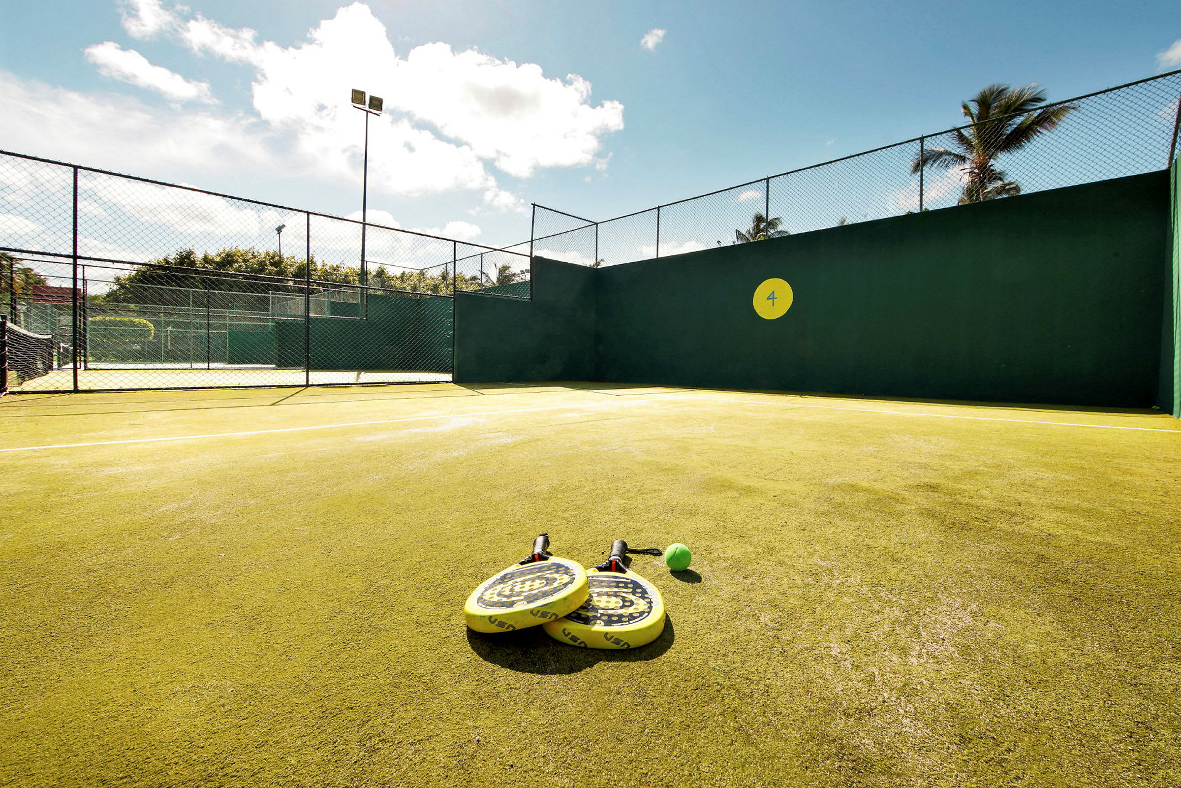Padelkenttä, jossa keltaiset mailat ja pallo maassa. Aurinkoinen sää. (Tekoälyn luoma teksti)
