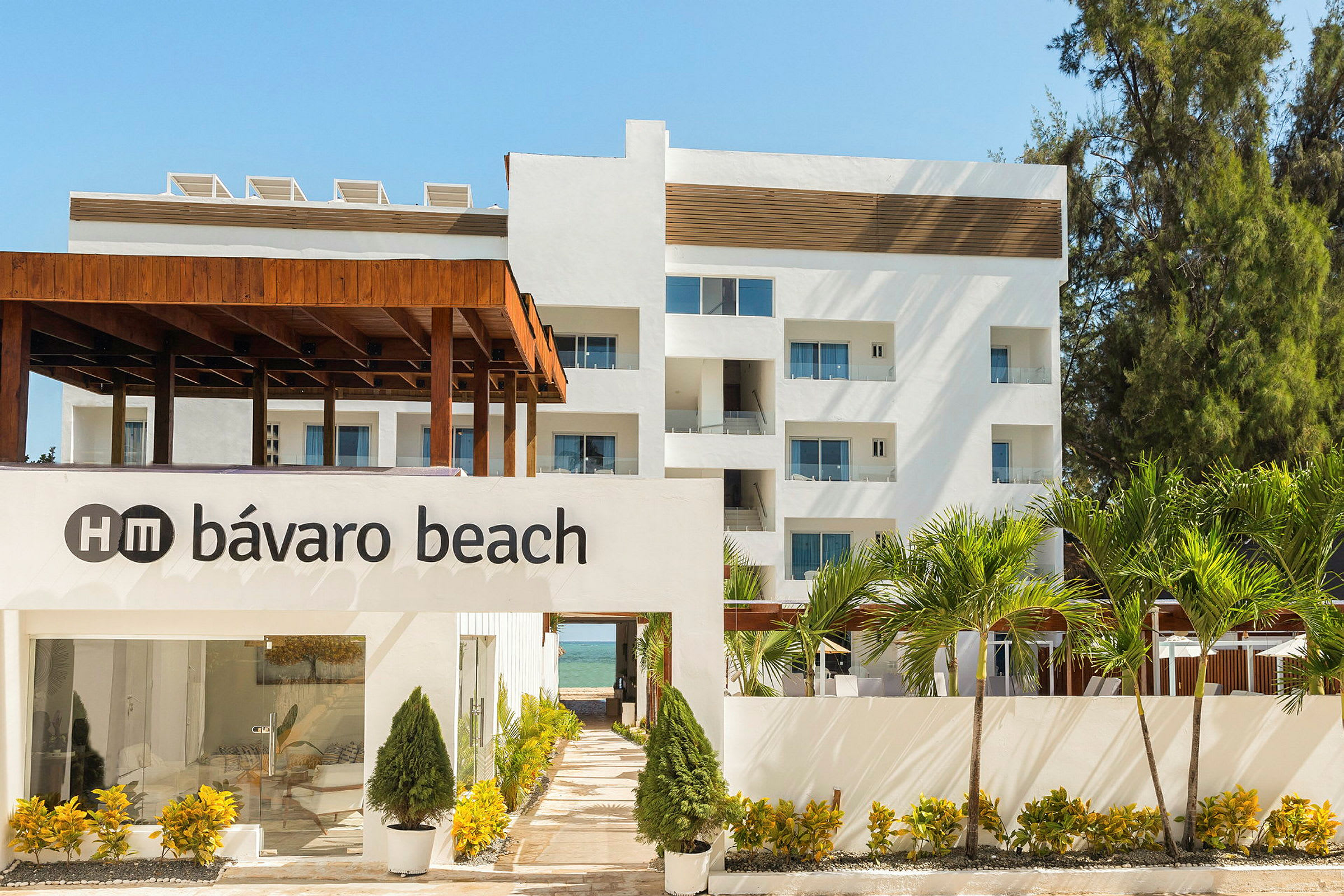 Kuva valkoisesta rakennuksesta, jonka edessä on seinässä teksti "HM Bávaro Beach", ympärillä vehreää kasvillisuutta ja palmuja. (Tekoälyn luoma teksti)