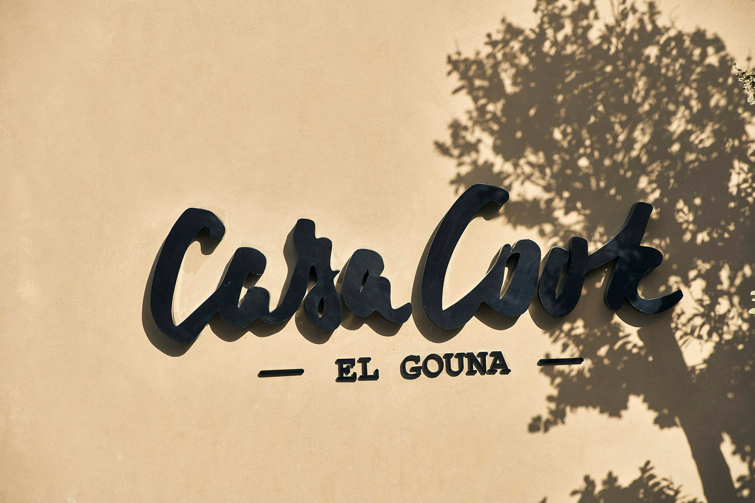 Casa Cook El Gounan logo seinällä, puun varjo. (Tekoälyn luoma teksti)