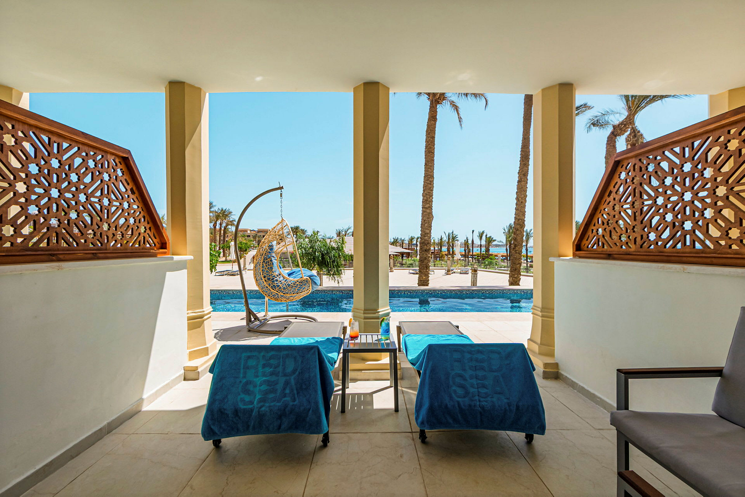 Kaksi aurinkotuolia, joissa on siniset kylpypyyhkeet, sijaitsee terassilla, jolta avautuu näkymä altaalle ja palmuihin. (Tekoälyn luoma teksti)