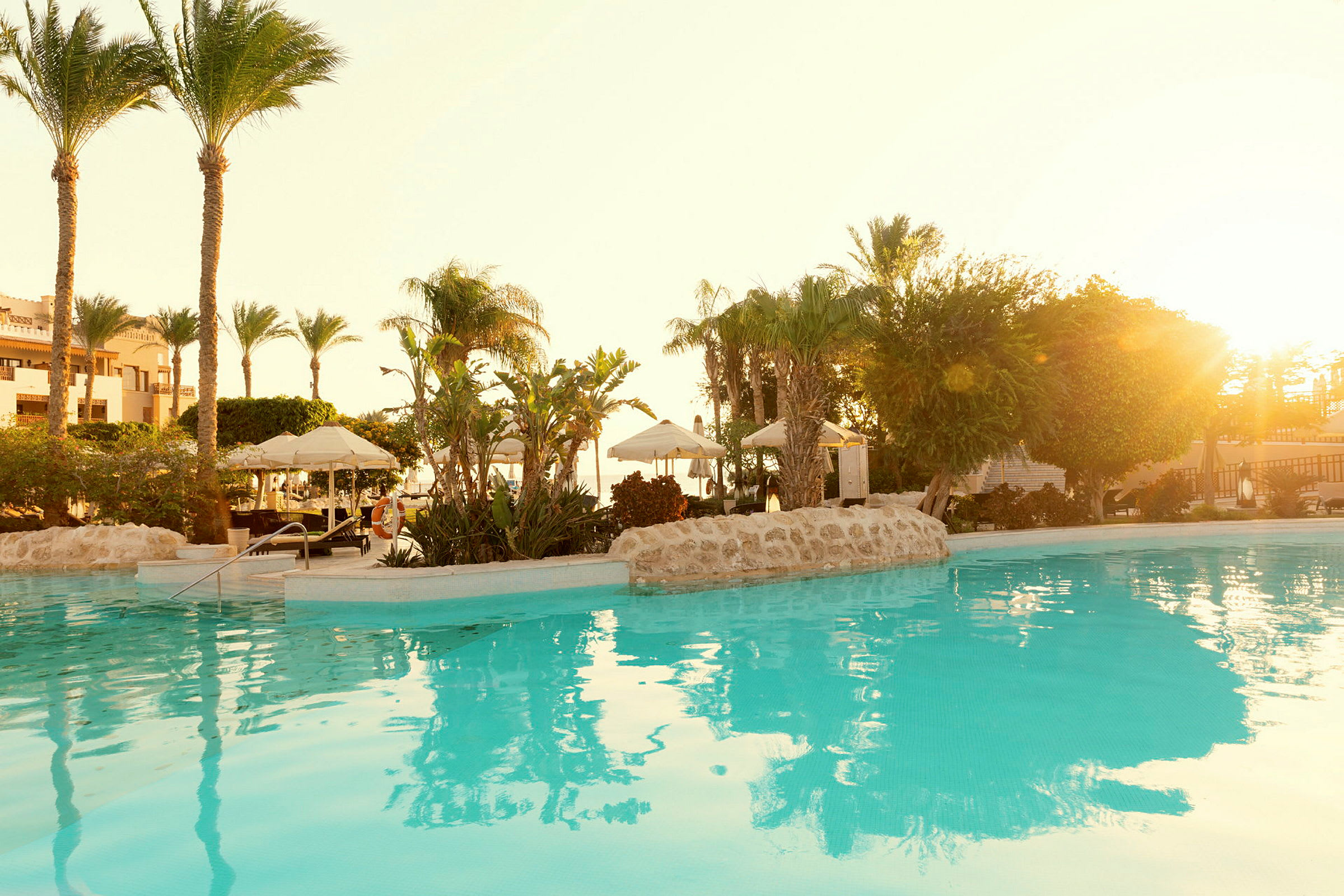 Aurinko valaisee uima-allasalueen, jossa on palmuja ja aurinkovarjoja veden äärellä. (Tekoälyn luoma teksti)