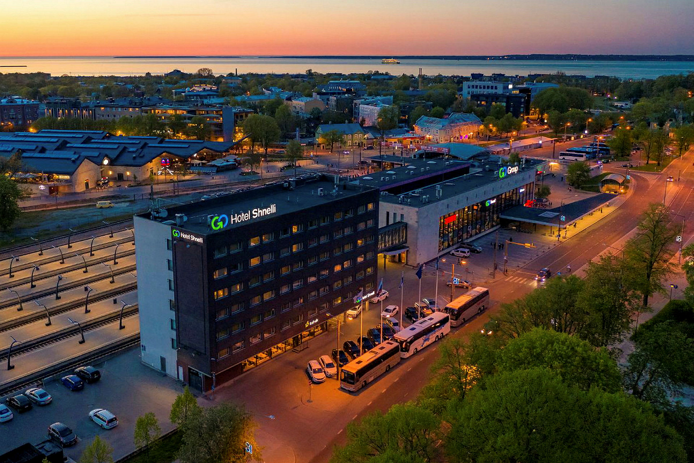 Suurempi rakennus, jonka kyltti "Hotel Shnelli" näkyy tien vieressä, jota ympäröivät bussit ja autot. Alue on valaistu illalla, ja taustalla näkyy puita ja satama. (Tekoälyn luoma teksti)