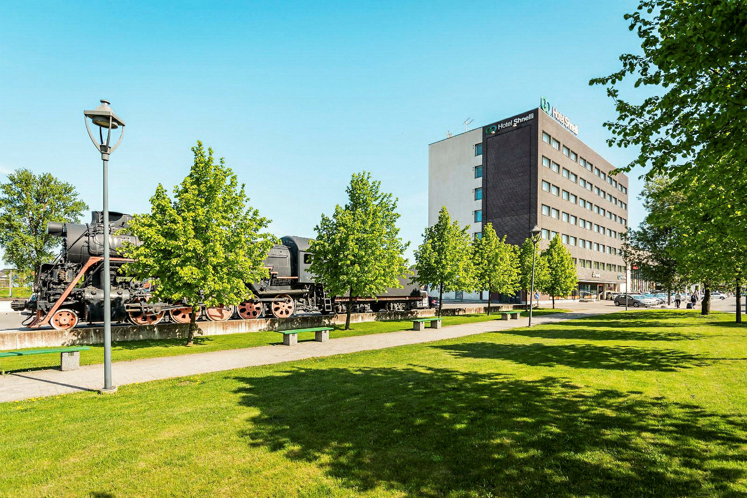 Vanha höyryveturi seisoo ulkona modernin hotellin vieressä vehreiden puiden keskellä. (Tekoälyn luoma teksti)