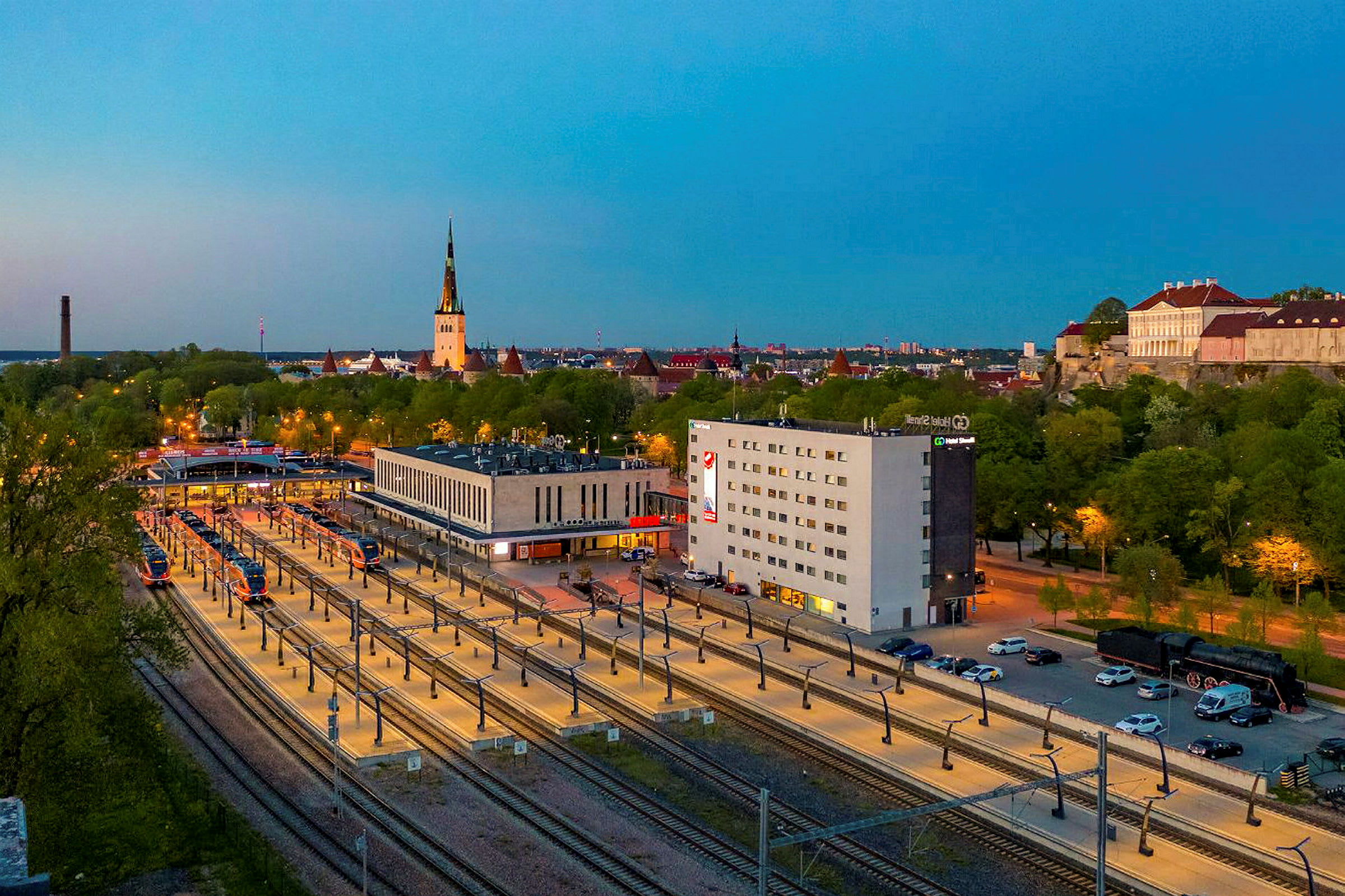 Kuva rautatieasemasta rakennusten ja vihreiden puiden ympäröimänä, taustalla näkyy kirkontorni. (Tekoälyn luoma teksti)
