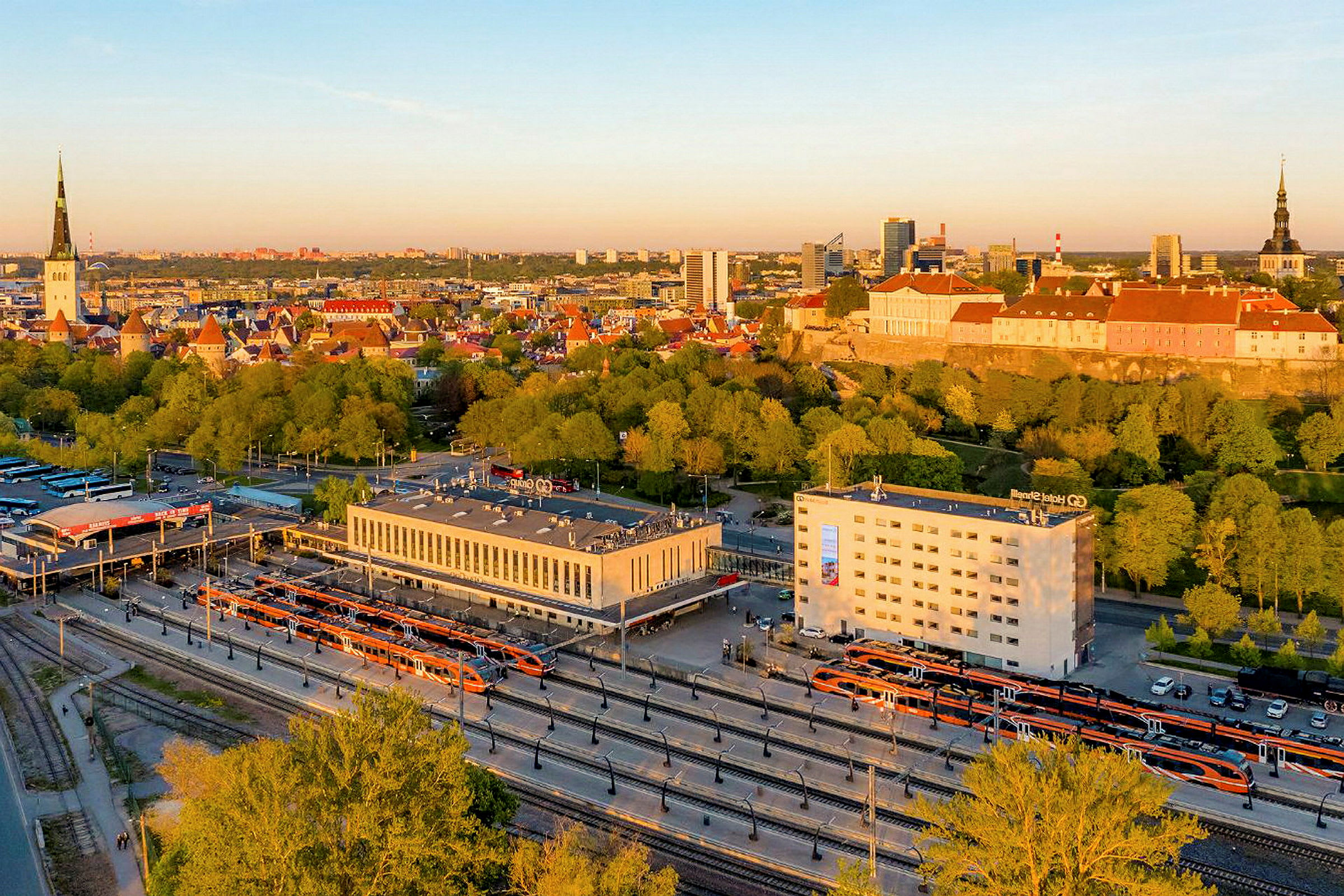 Kuva kaupungista, jonka etualalla on rautatieasema ja taustalla kaupungin siluetti rakennuksineen ja puineen. (Tekoälyn luoma teksti)