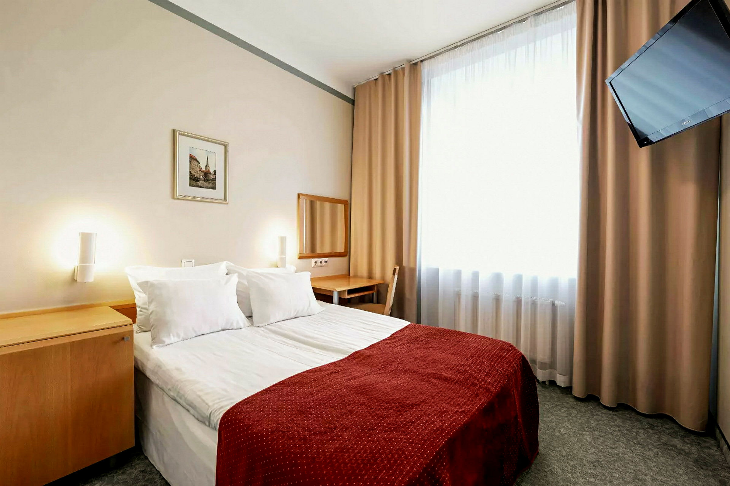 Hotellihuone, jossa on parivuode, punainen sängynpäällinen. Seinän vierellä on peili ja kirjoituspöytä. Huoneessa on ikkuna, jossa on vaaleat verhot ja seinään kiinnitetty televisio. (Tekoälyn luoma teksti)