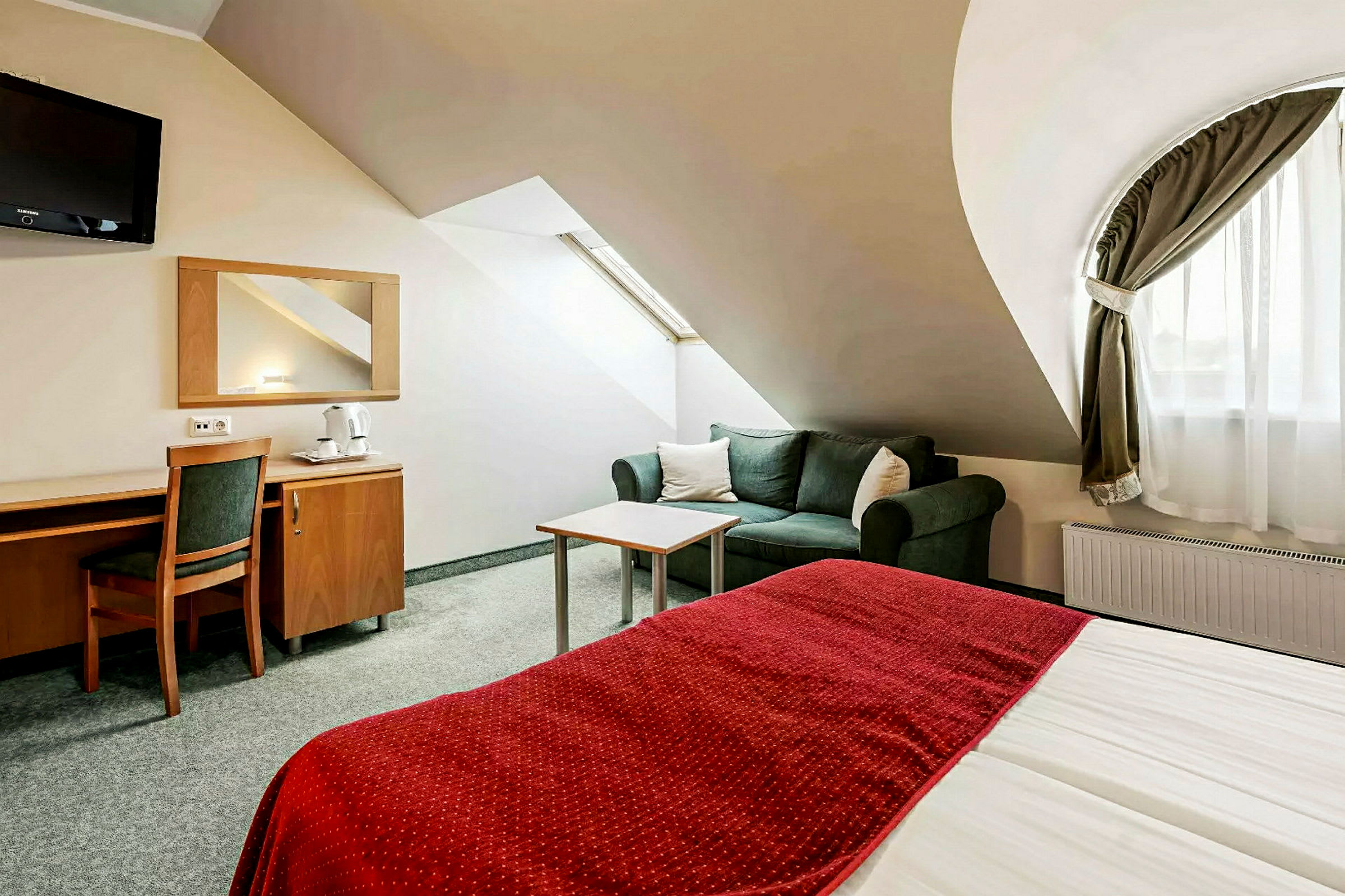 Hotellihuone, jossa on punainen peite, sohva ja kirjoituspöytä tuoleineen. (Tekoälyn luoma teksti)
