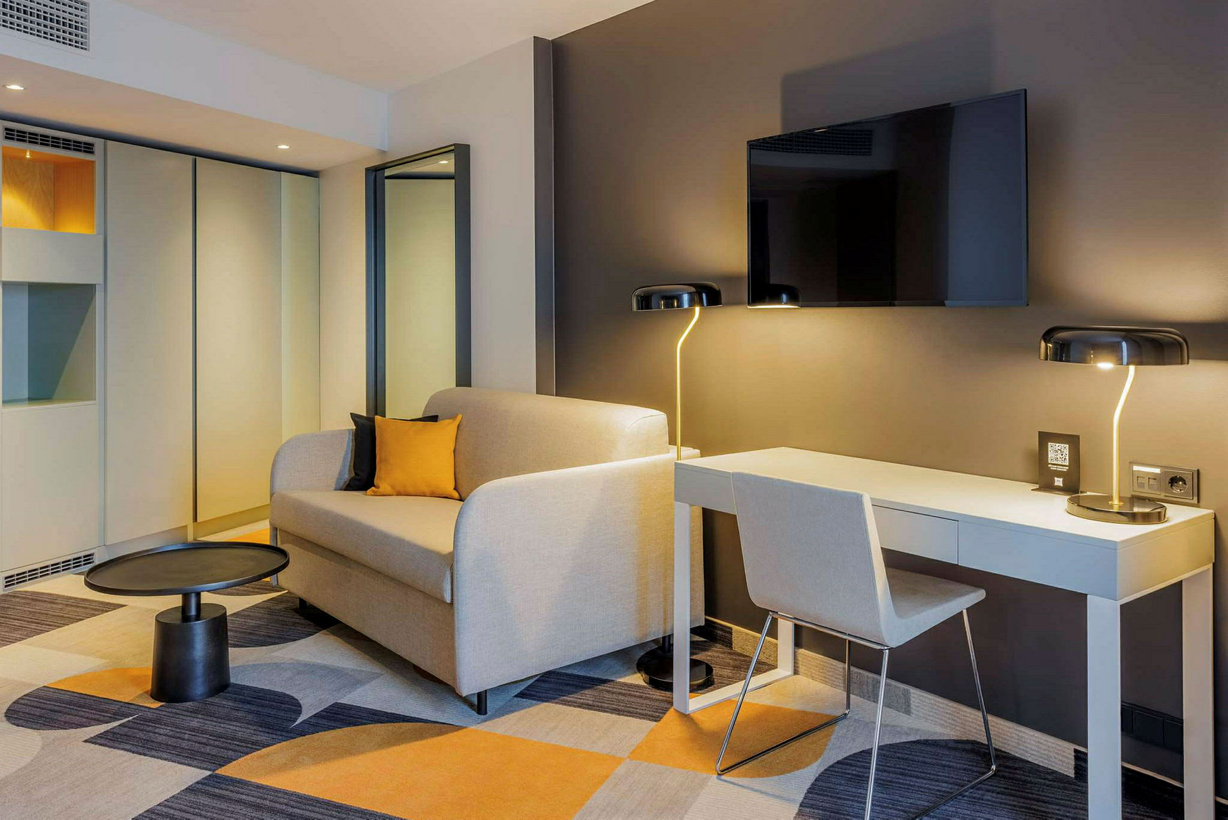 Moderni hotellihuone, jossa on sohva, kirjoituspöytä ja seinään kiinnitetty TV. (Tekoälyn luoma teksti)