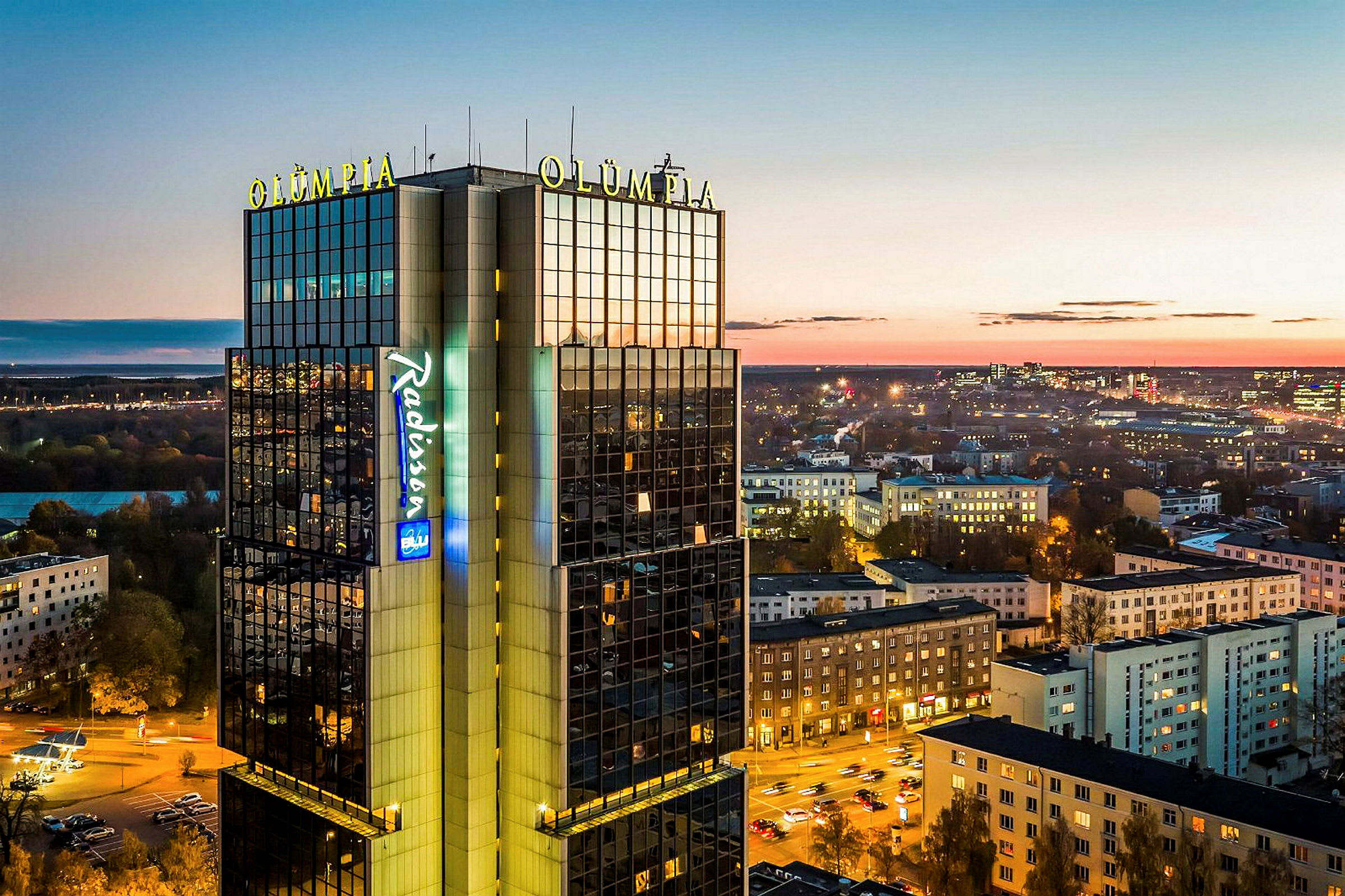 Korkea rakennus, jossa lukee "Olümpia" ja "Radisson", kaupunkinäkymä hämärässä. (Tekoälyn luoma teksti)