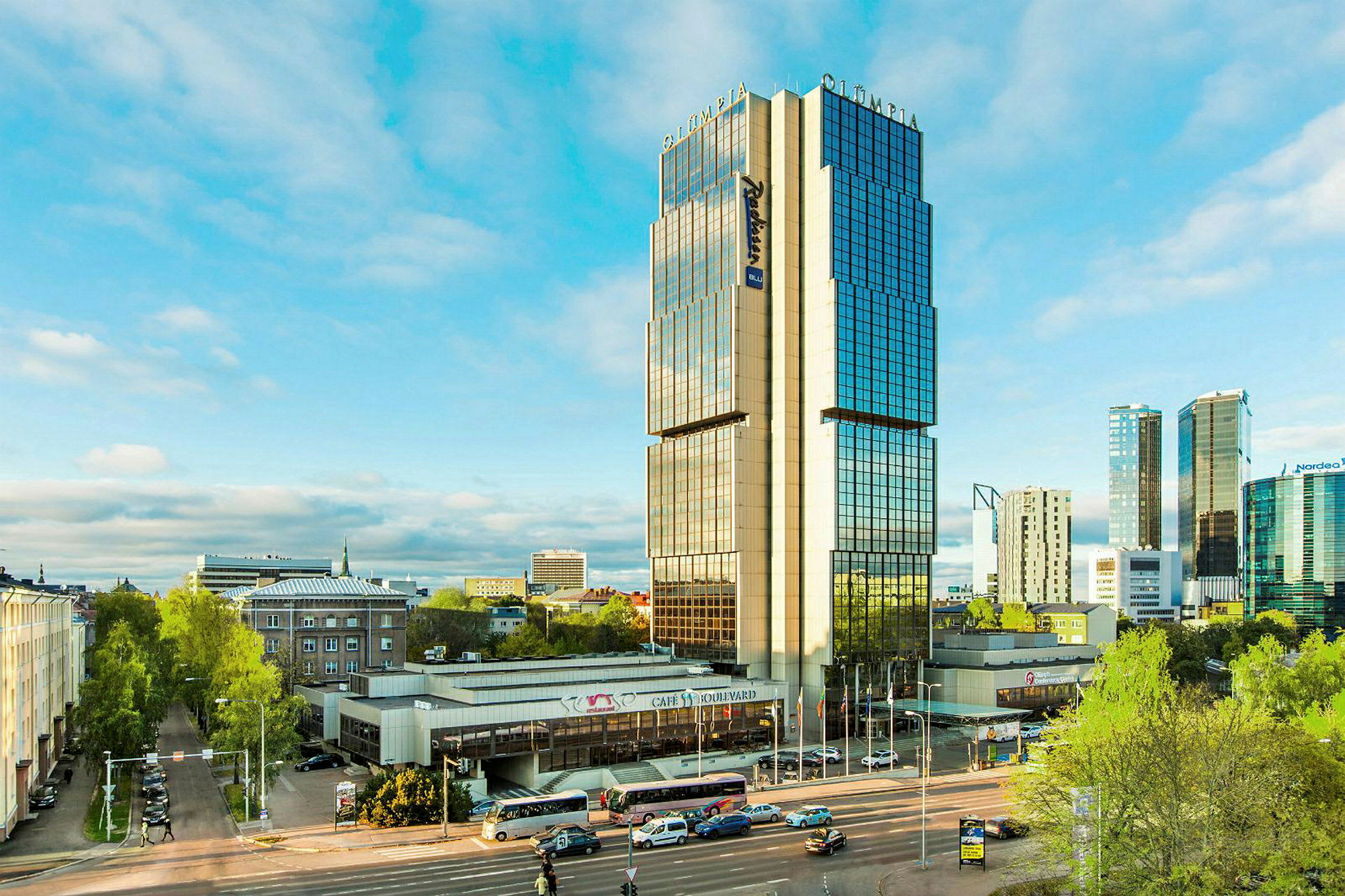 Kaupunkimaisema, jossa on moderni korkea rakennus, jossa on heijastavia ikkunoita. Rakennuksen edessä on tie, jolla on useita ajoneuvoja. (Tekoälyn luoma teksti)