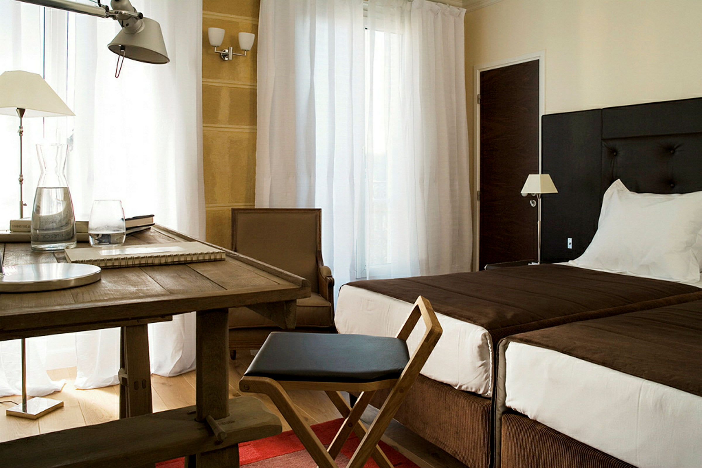Hotellihuone, jossa kaksi vuodetta, pöytä ja tuoli sekä nojatuoli valkoisten verhojen edessä. (Tekoälyn luoma teksti)