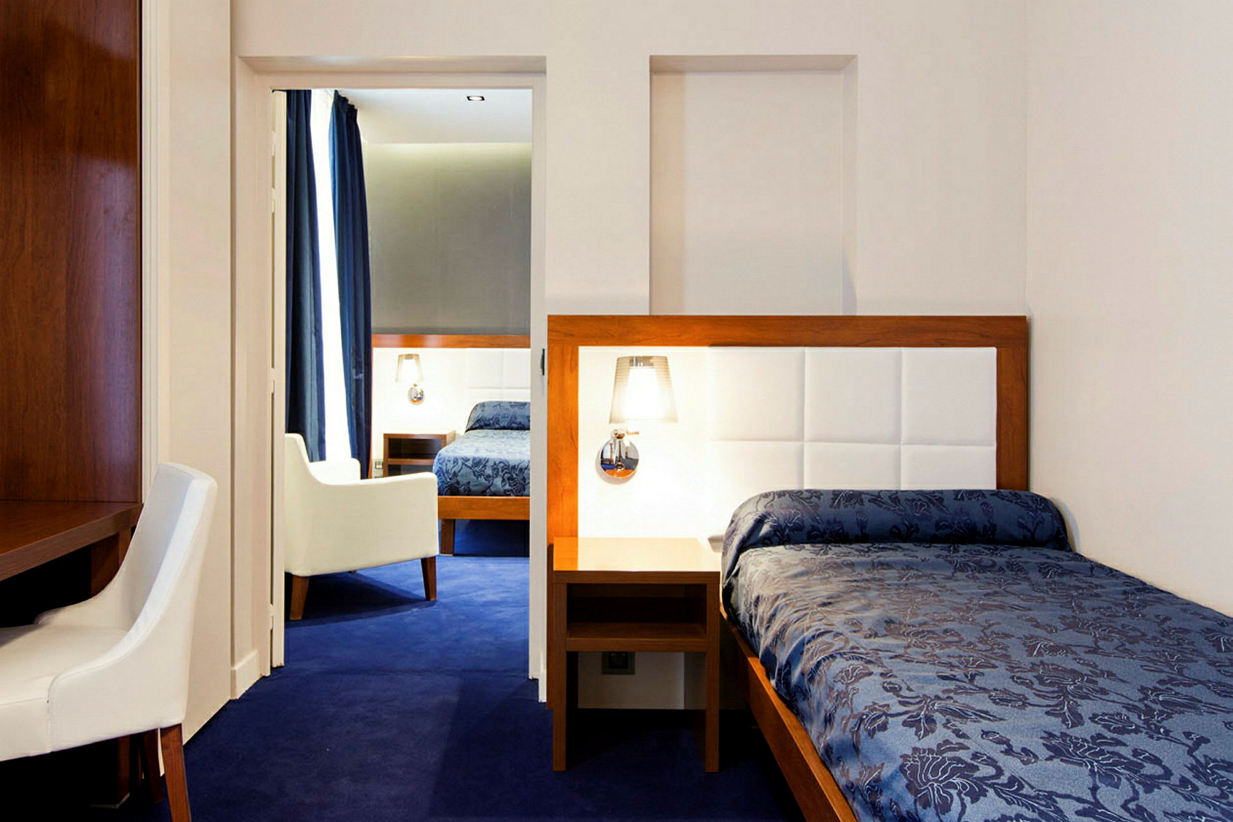 Hotellihuone, jossa sininen matto, yhden hengen sänky sinisellä päiväpeitteellä ja vaaleat huonekalut. (Tekoälyn luoma teksti)