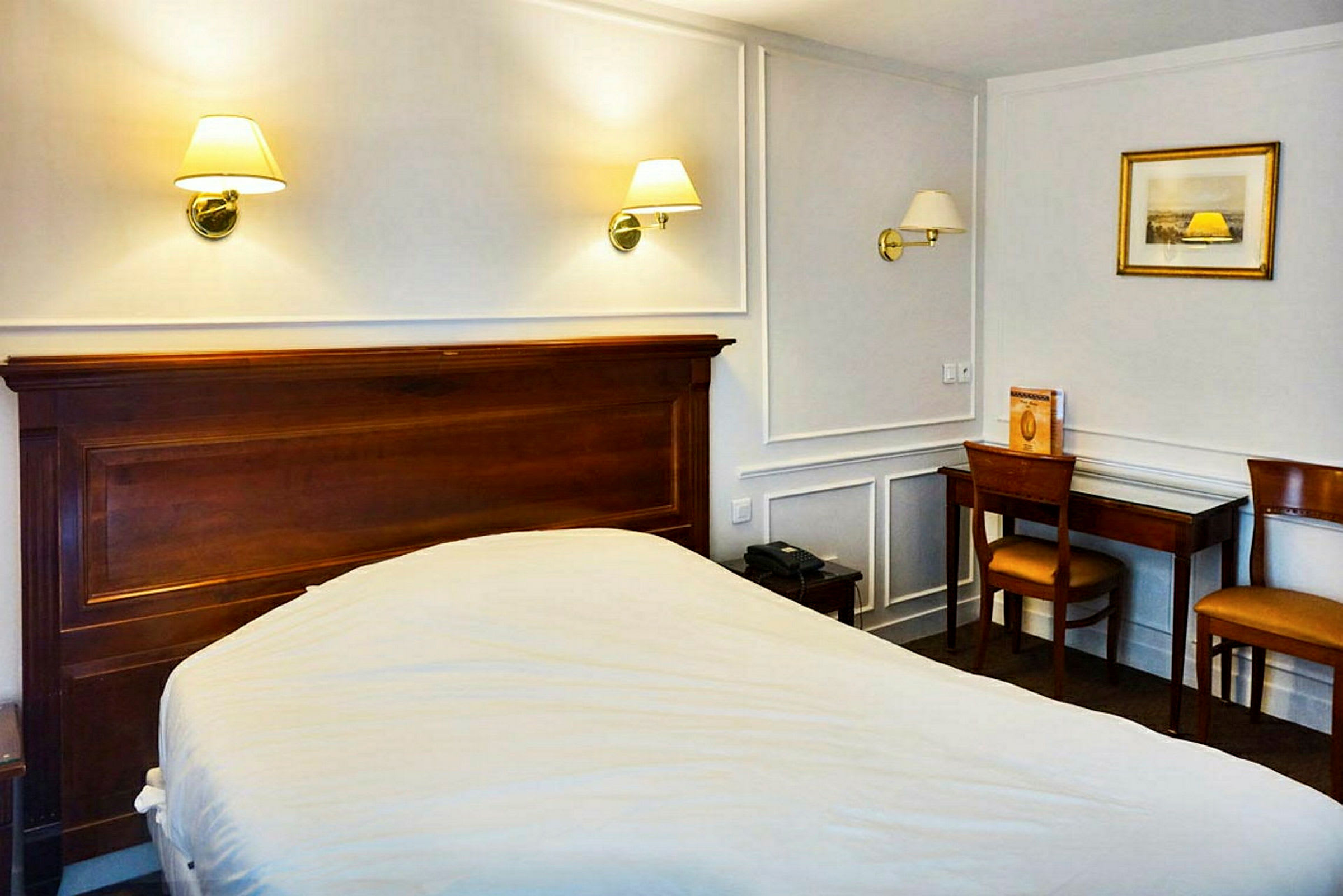 Hotellihuone, jossa on sänky, puupöytä ja tuoleja sekä seinävalaisimia sängyn yläpuolella. (Tekoälyn luoma teksti)