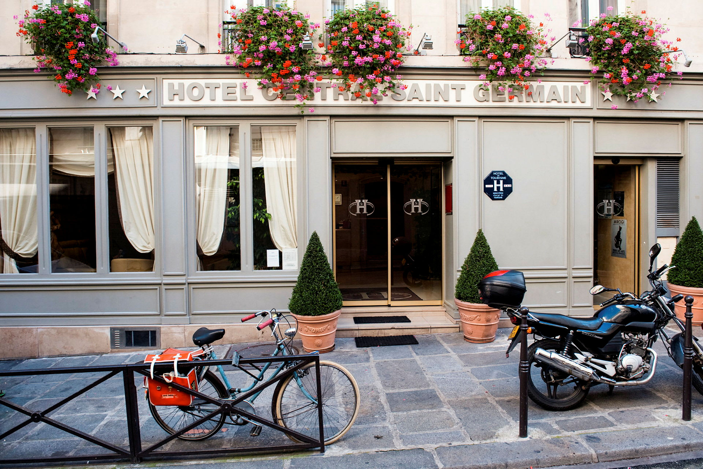 Hotellin julkisivu, jossa kyltti 'Hotel Central Saint Germain', kukkaruukkuja oven yläpuolella sekä polkupyörä ja moottoripyörä pysäköity edessä. (Tekoälyn luoma teksti)