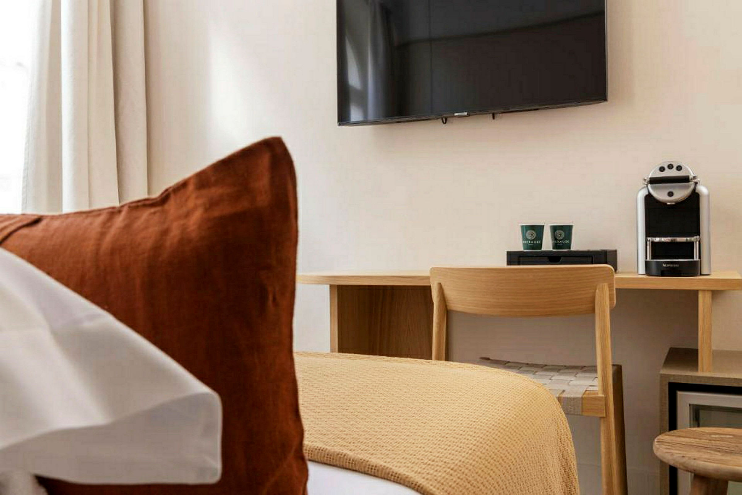 Hotellihuone, jossa on sänky etualalla, työpöytä ja tuoli, kahvinkeitin sekä seinälle kiinnitetty televisio. (Tekoälyn luoma teksti)