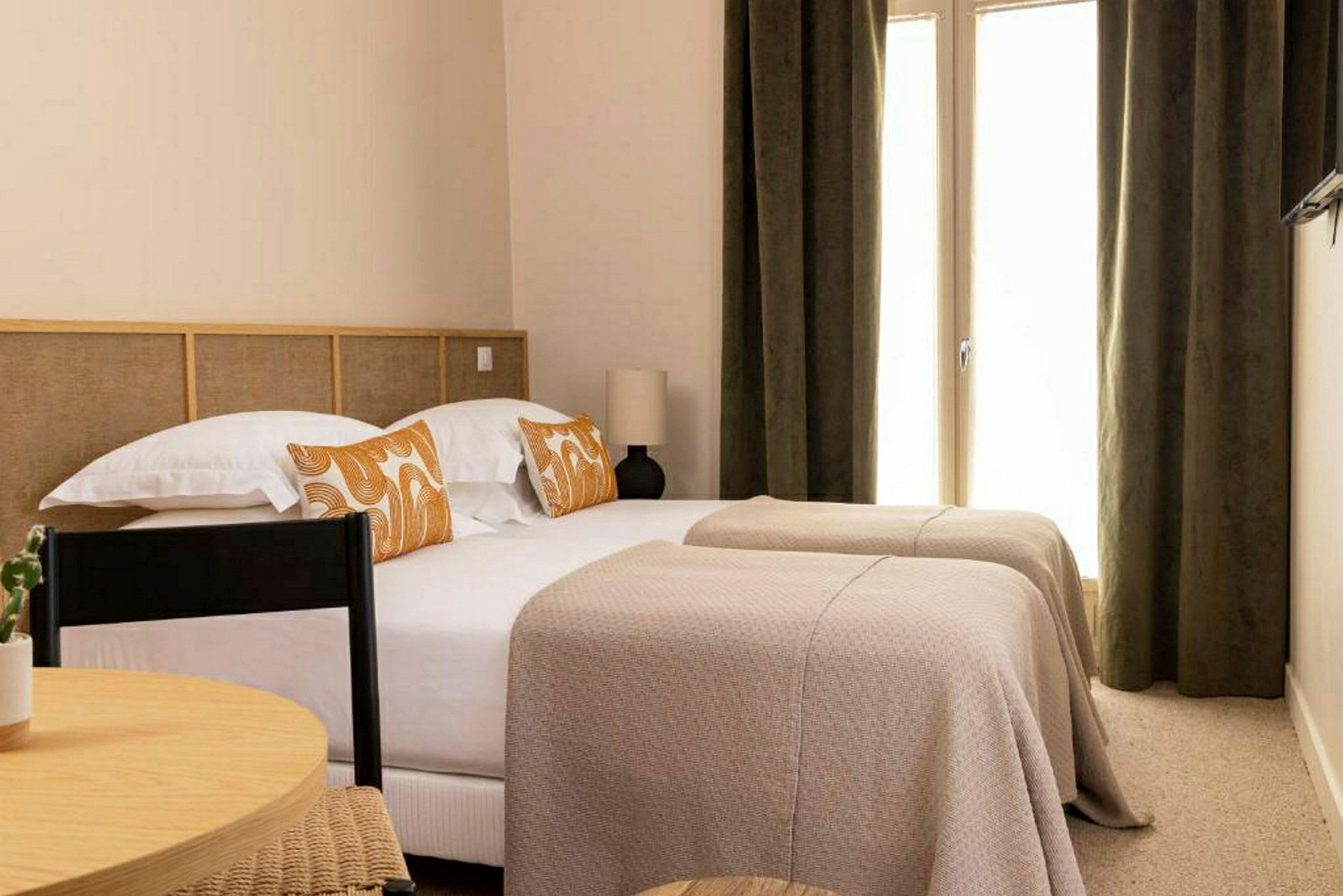 Hotellihuone, jossa on kaksi yksittäistä sänkyä, päällystetty beigeillä päiväpeitteillä ja koristeltu oranssikuviollisilla tyynyillä. (Tekoälyn luoma teksti)
