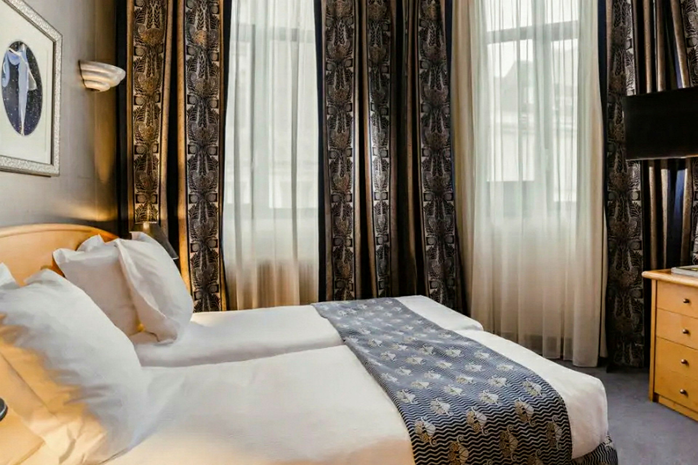 Hotellihuone, jossa kaksi erillistä vuodetta, vaaleat verhot ja koristeellinen sängynpäällinen. (Tekoälyn luoma teksti)