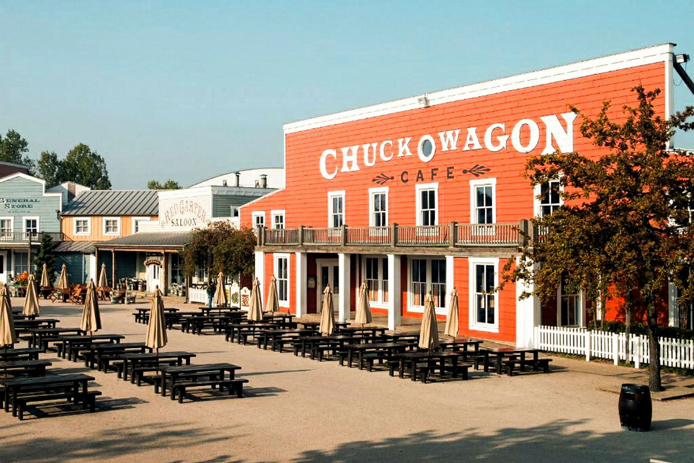 Alue, jolla on pöytiä ja aurinkovarjoja rakennuksen edessä, jossa lukee 'Chuck Wagon Cafe'. (Tekoälyn luoma teksti)