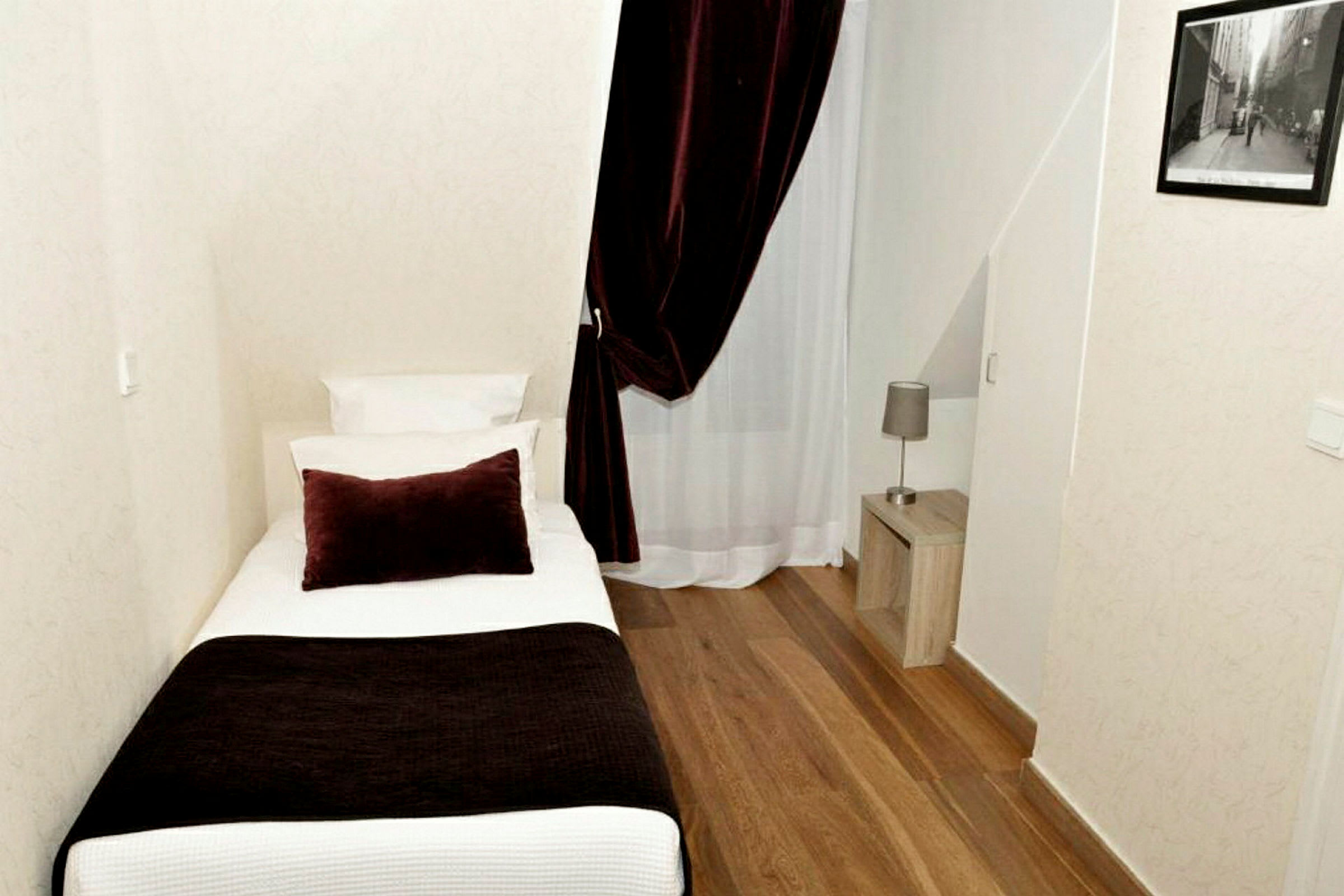 Hotellihuone, jossa on yksittäinen sänky, yöpöytä ja lamppu. Huoneessa on puulattia ja verho ikkunan edessä. (Tekoälyn luoma teksti)