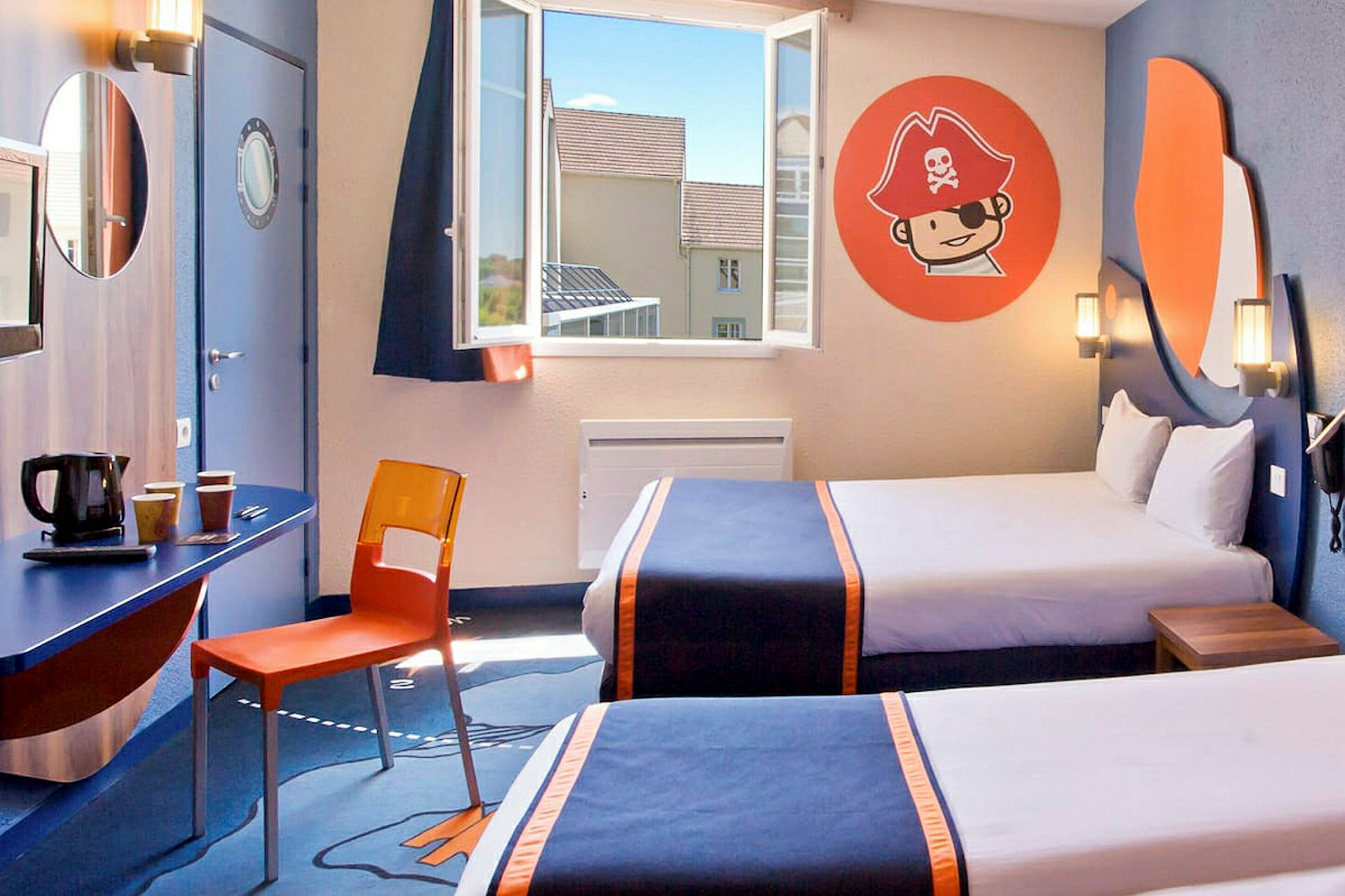 Hotellihuone, jossa on kaksi sänkyä, siniset päiväpeitot ja oranssit yksityiskohdat. Ikkuna on avoin ja huoneessa on merirosvoteemainen seinäkoriste, sekä kirjoituspöytä, jolla on oranssi tuoli ja kahvinkeitin pyöreän peilin alla. (Tekoälyn luoma teksti)