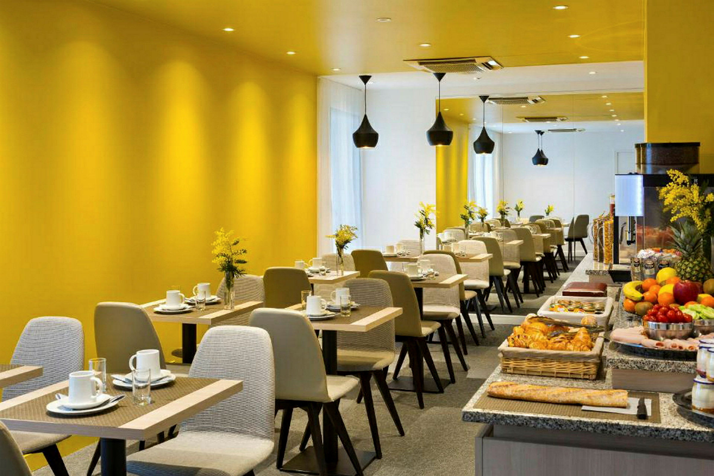 Moderni ravintola-alue, jossa on keltaiset seinät, katettuja pöytiä ja seisova pöytä erilaisine ruokalajeineen. (Tekoälyn luoma teksti)