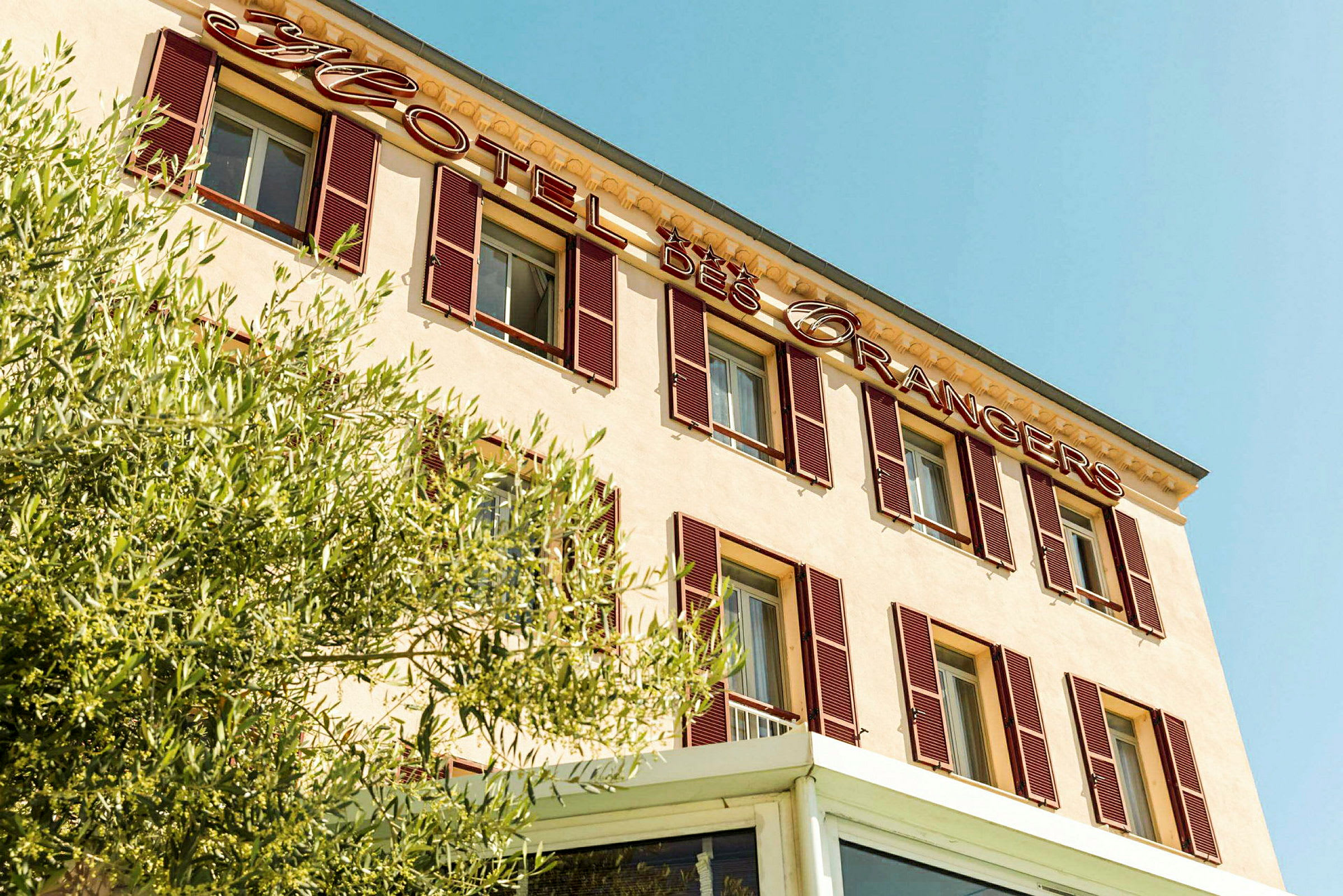 Hotellirakennuksen julkisivu, jossa on 'Hotel des Orangers' -kyltti ja kasvillisuutta etualalla. (Tekoälyn luoma teksti)