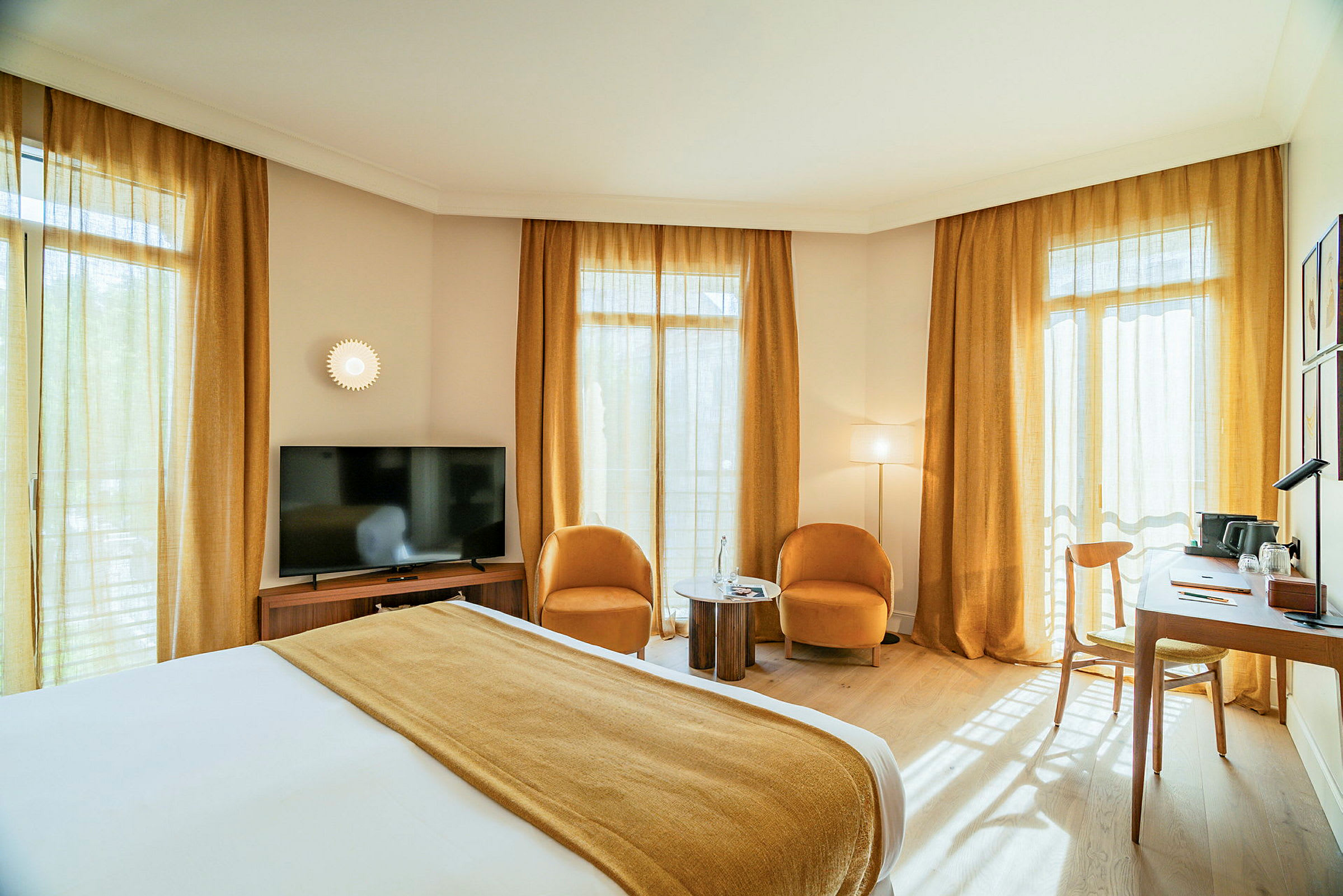 Hotellihuone, jossa on parivuode, kaksi nojatuolia ja pieni pöytä, televisio ja työpöytä ikkunoiden ääressä. (Tekoälyn luoma teksti)