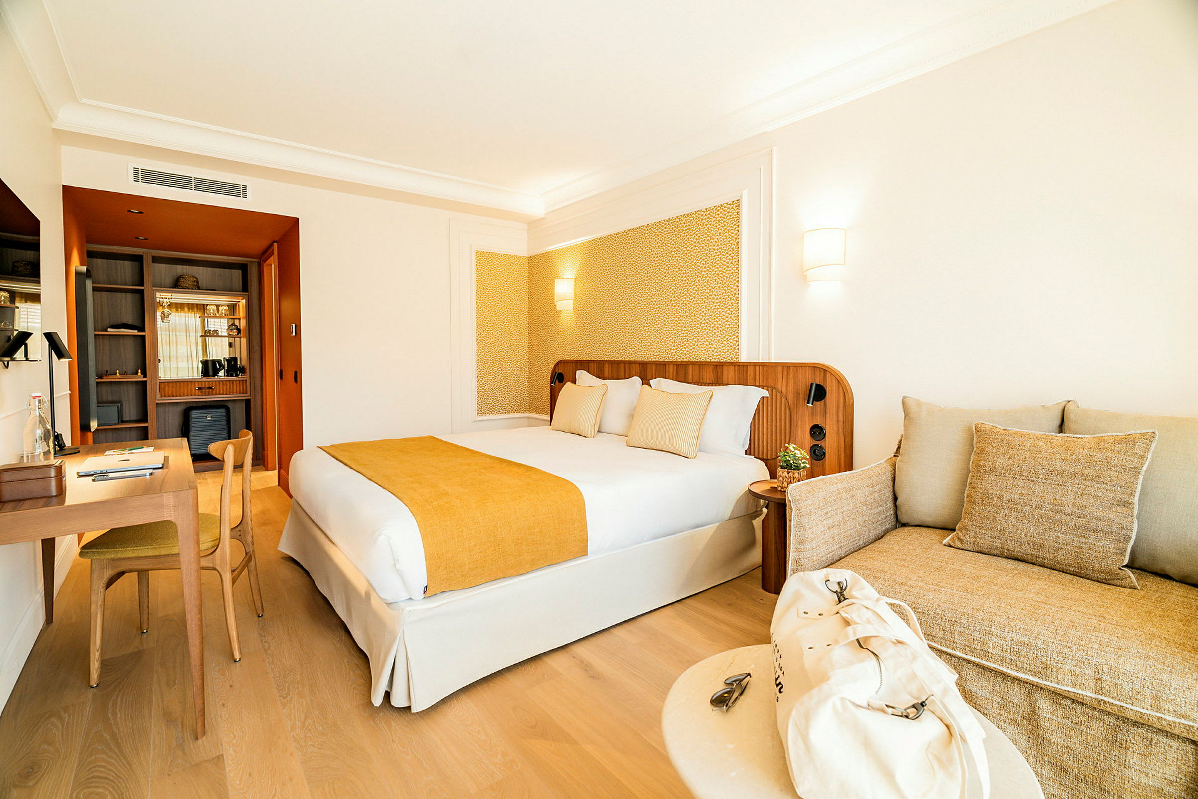 Hotellihuoneen sisustus, jossa on parivuode keltaisella päiväpeitolla ja tyynyillä, kirjoituspöytä tuoleineen vasemmalla ja beige tyynyillä varustettu sohva oikealla. (Tekoälyn luoma teksti)