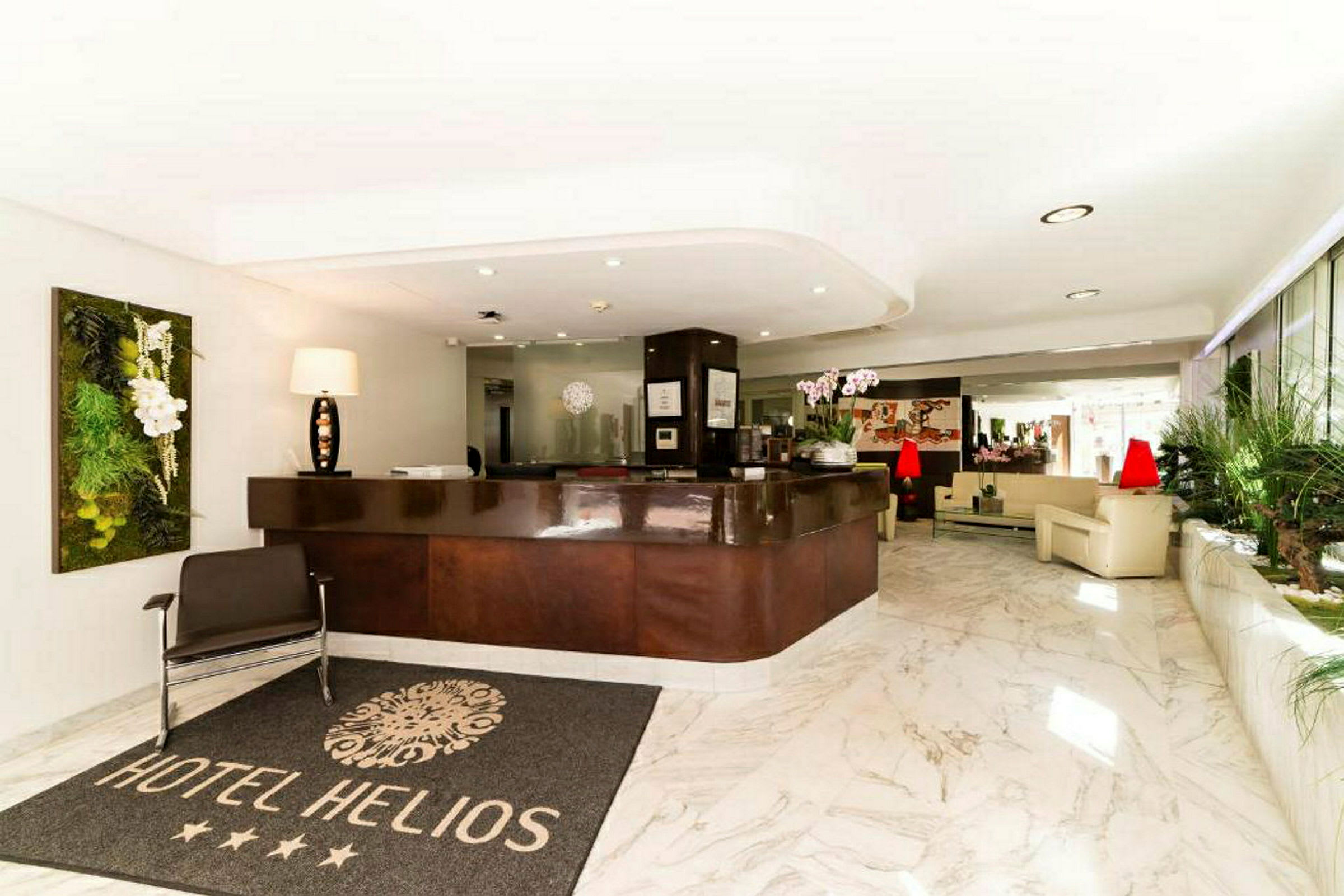 Hotellin aula, jossa vastaanotto, matto, jossa teksti "Hotel Helios", ja istuimia valoisassa ympäristössä. (Tekoälyn luoma teksti)