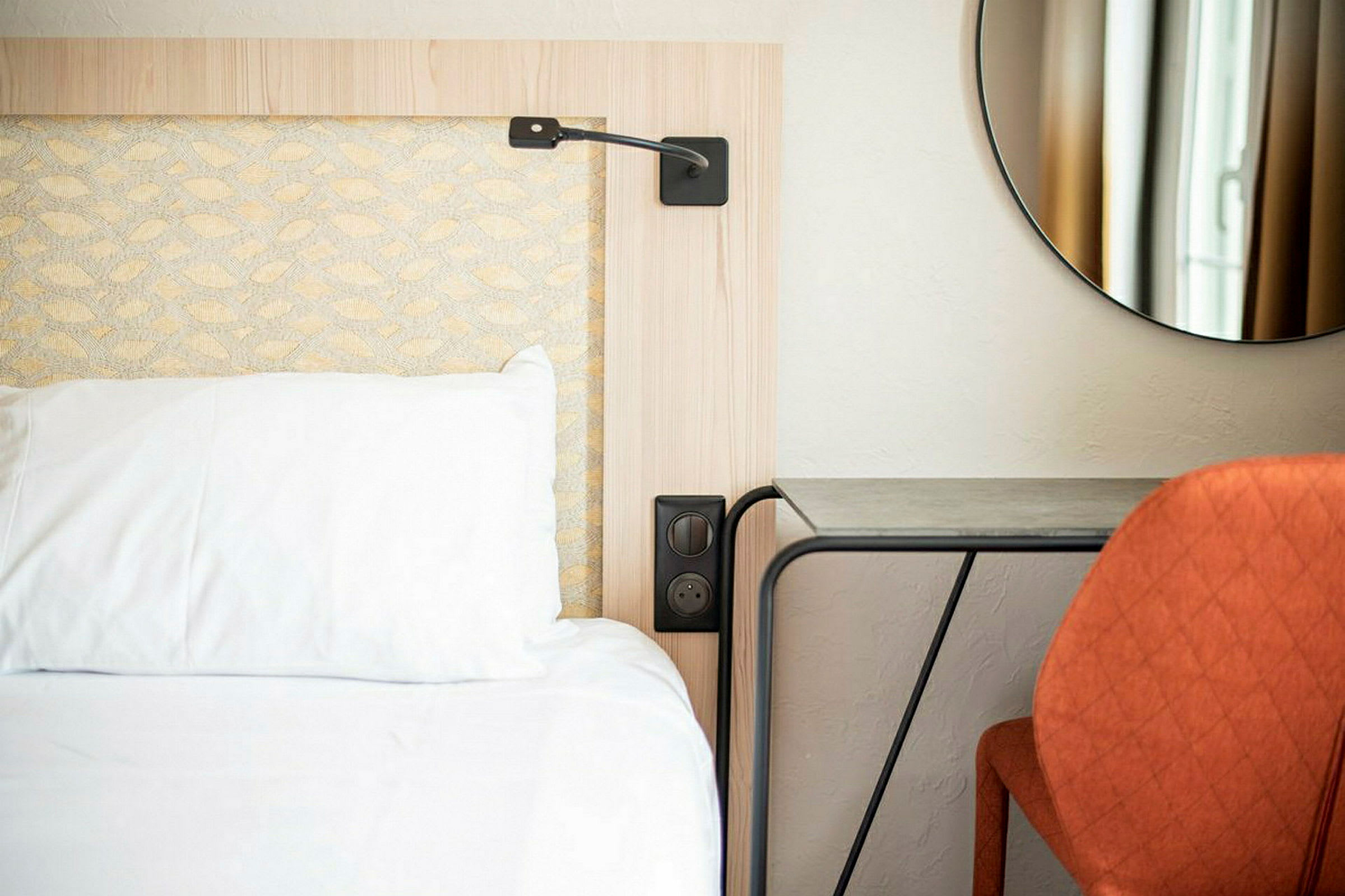 Hotellihuoneen sänky, jossa on valkoinen tyyny ja yöpöytä vieressä, sekä oranssi tuoli pöydän edessä ja pyöreä peili seinällä. (Tekoälyn luoma teksti)