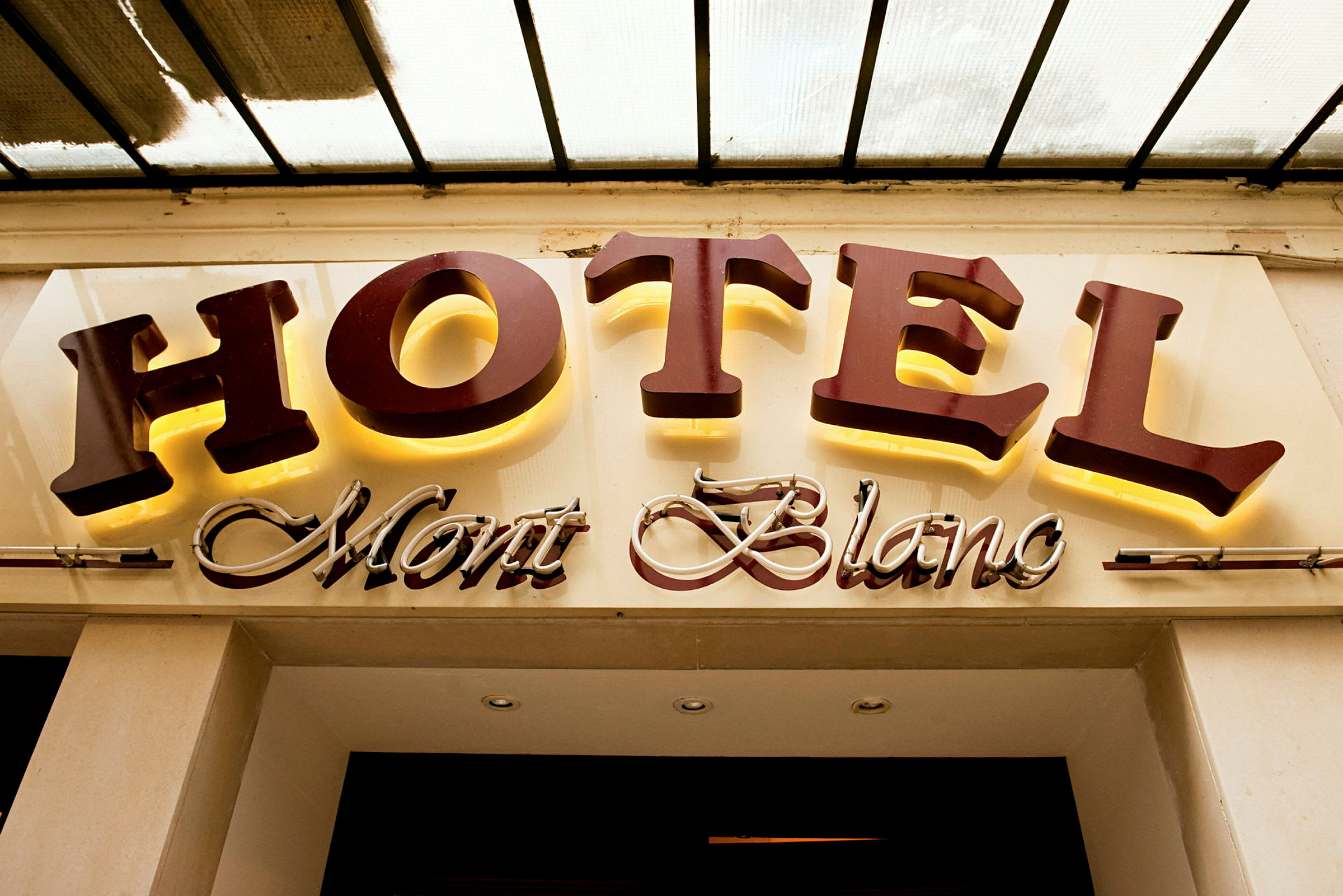 Hotellin kyltti, jossa lukee "HOTEL Mont Blanc"; "HOTEL" on suurilla, valaistuilla kirjaimilla ja "Mont Blanc" on kursiivilla koristeellisessa tyylissä. (Tekoälyn luoma teksti)