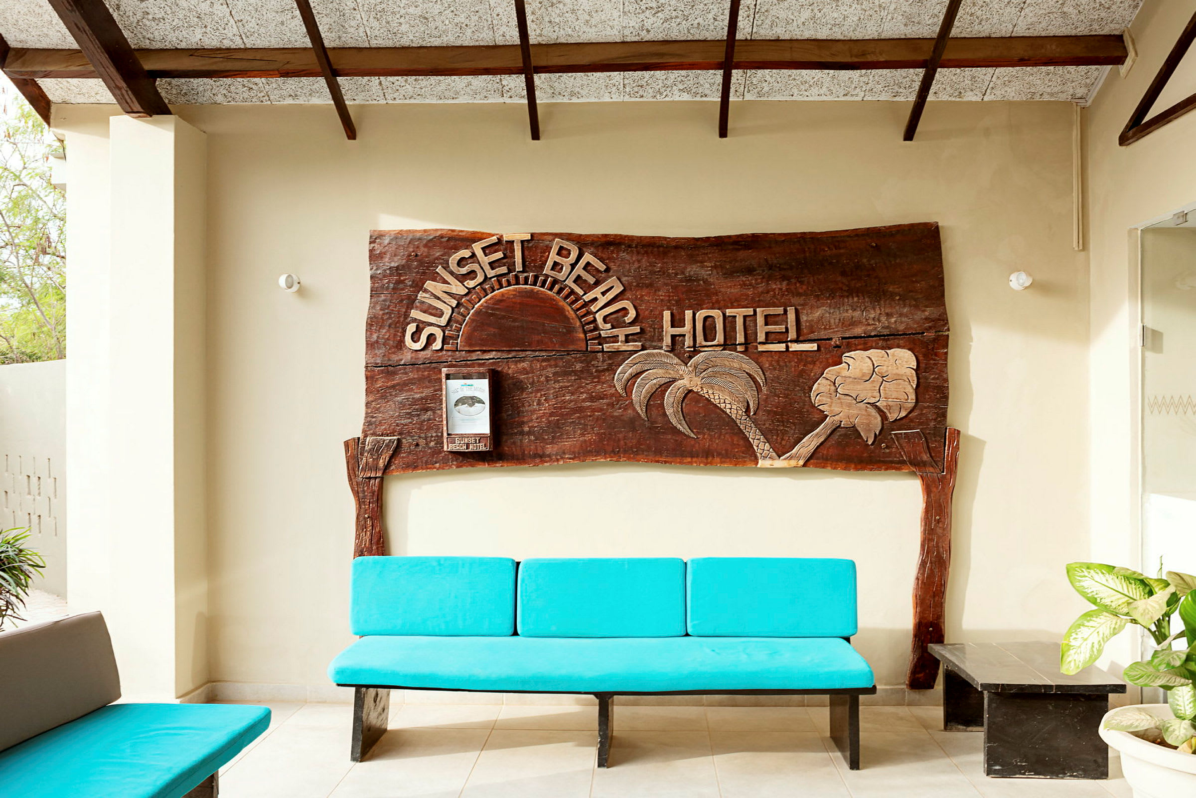 Seinässä on puinen kyltti, jossa lukee "Sunset Beach Hotel". Edessä on sininen penkki ja muutamia kasveja. (Tekoälyn luoma teksti)