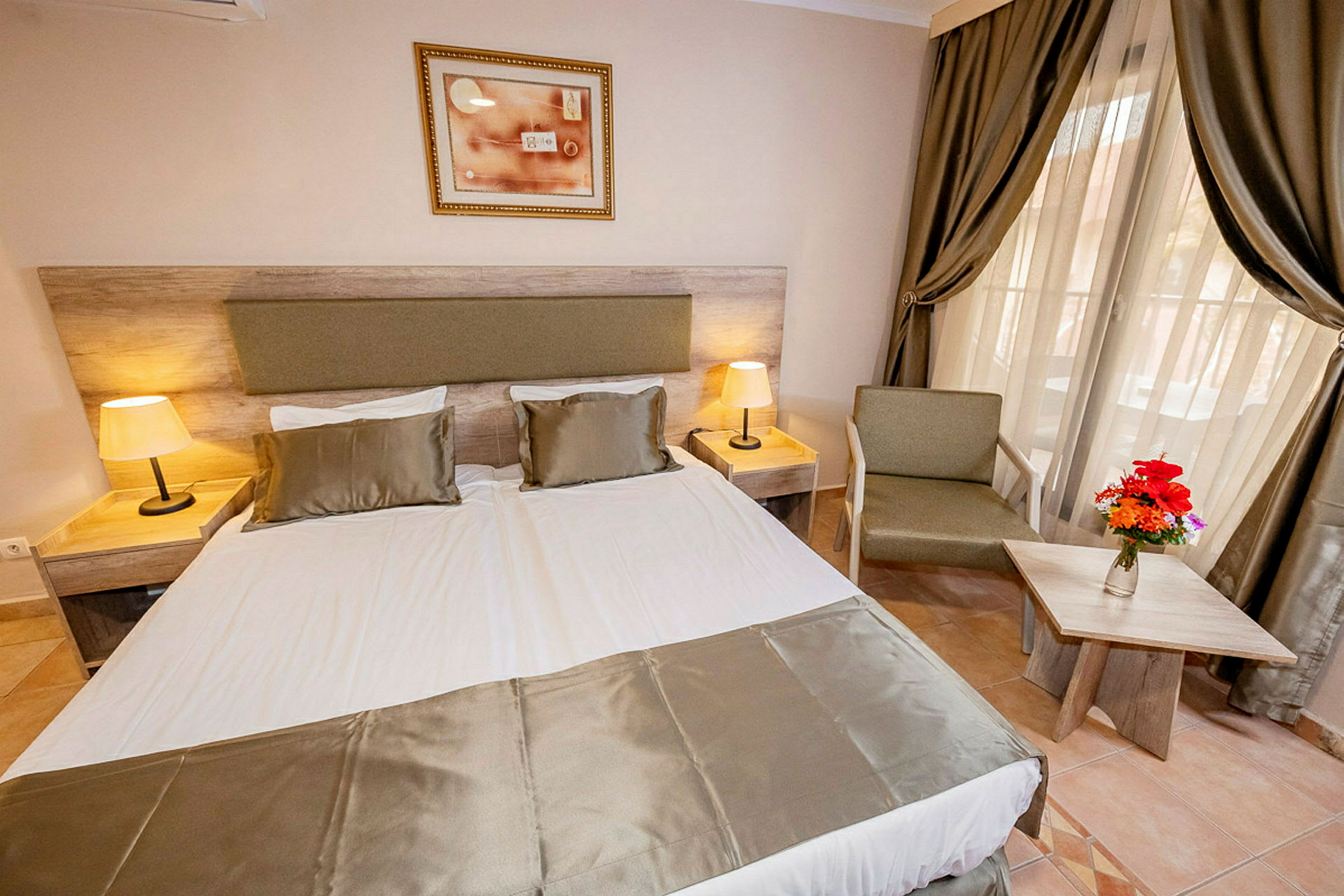 Hotellihuone, jossa on parisänky, kaksi yöpöytää lampuilla, nojatuoli ja pieni pöytä kukkavaasilla. (Tekoälyn luoma teksti)
