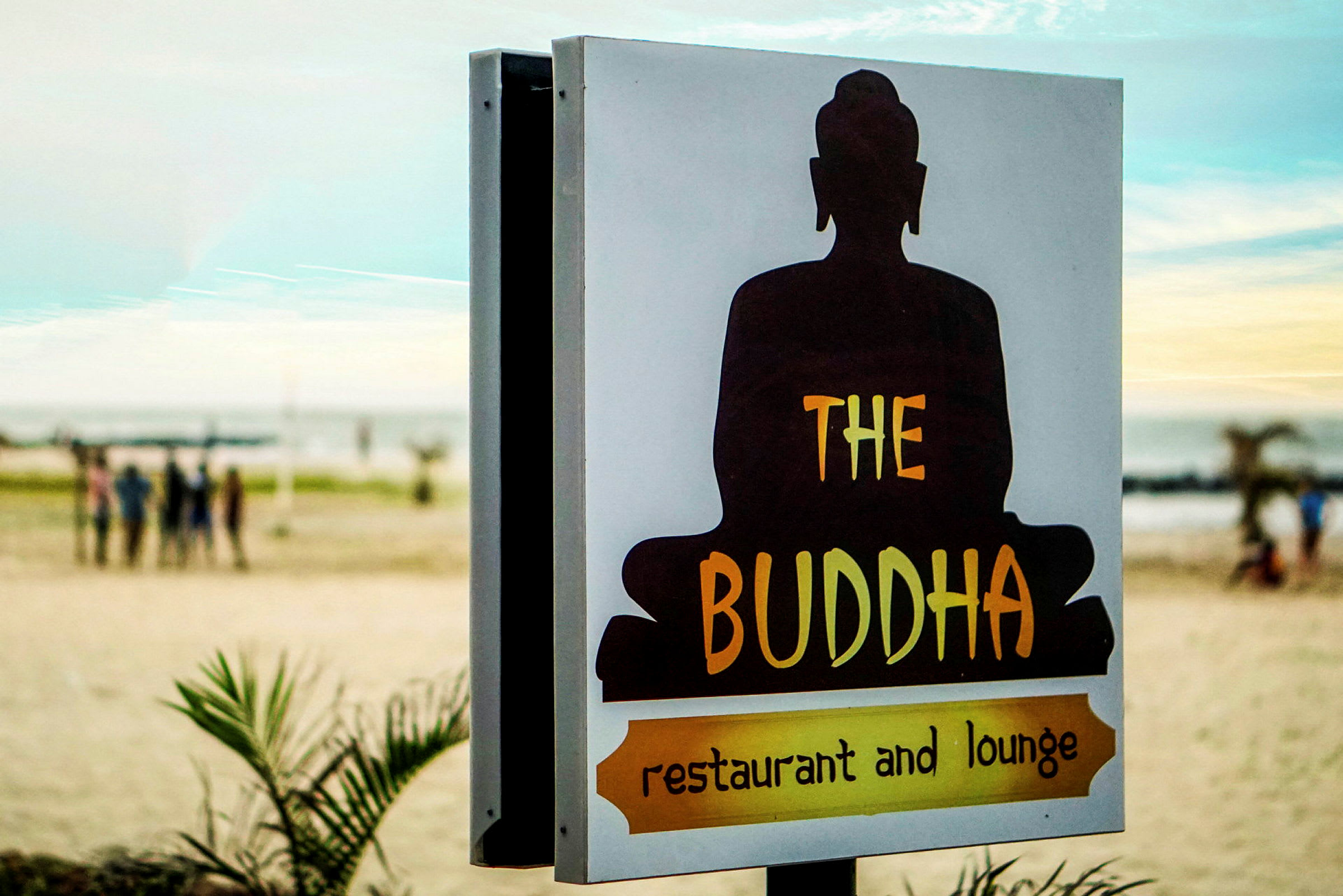 Kyltti, jossa lukee 'The Buddha restaurant and lounge' ja taustalla istuvan Buddhan siluetti sekä ranta. (Tekoälyn luoma teksti)