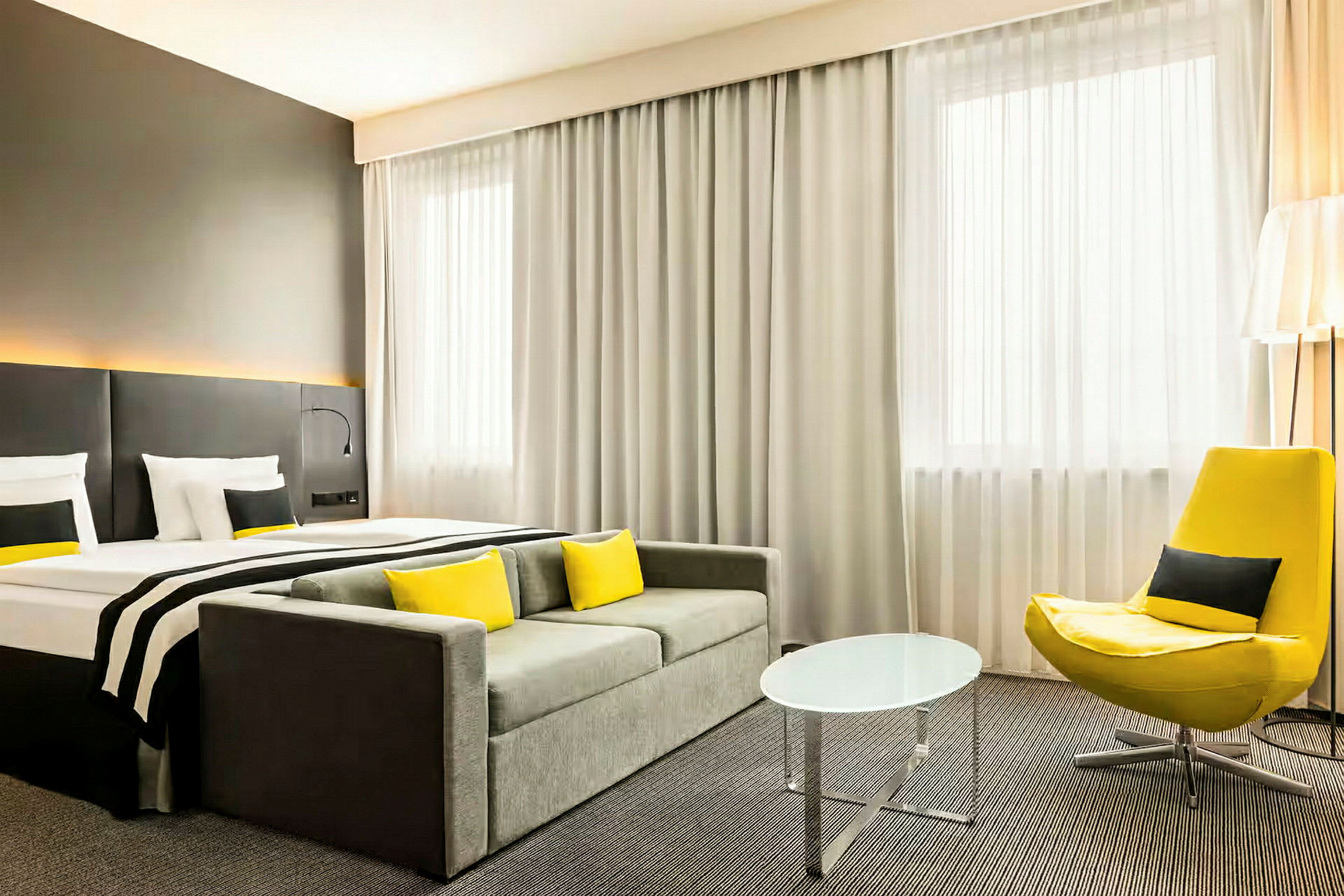 Hotellihuone, jossa on sänky, sohva, keltainen nojatuoli ja pieni pyöreä pöytä. (Tekoälyn luoma teksti)