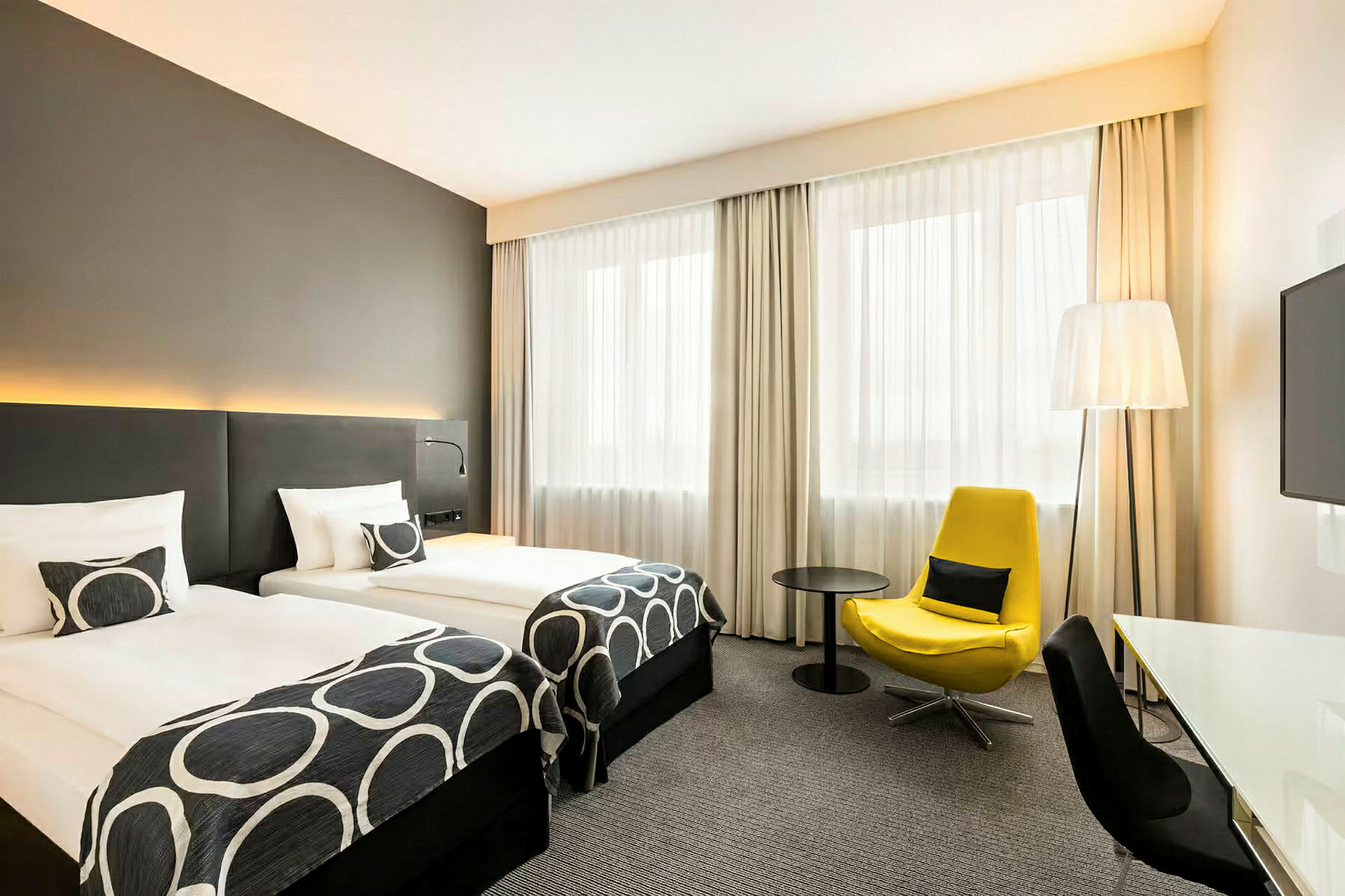 Hotellihuone, jossa on kaksi yhden hengen vuodetta, keltainen nojatuoli ja kirjoituspöytä. (Tekoälyn luoma teksti)
