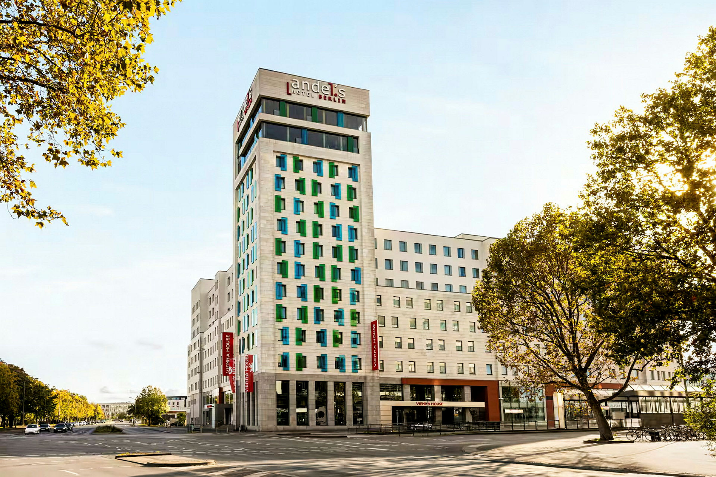 Moderni hotellirakennus, jossa on valkoinen julkisivu ja värikkäät ikkunat, ympärillä teitä ja puita. (Tekoälyn luoma teksti)