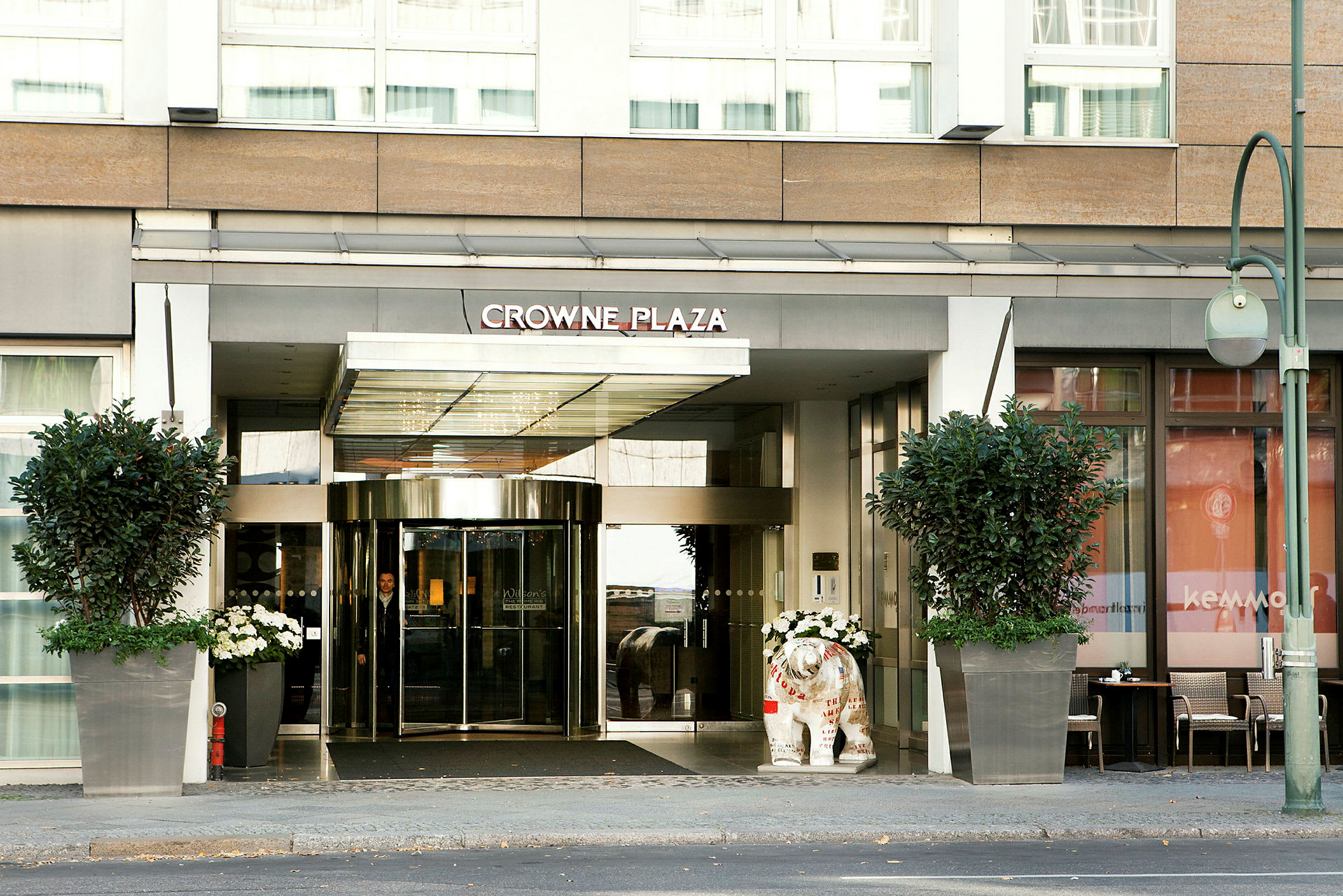 Crowne Plaza -hotellin sisäänkäynti lasiovilla ja suurilla kukkaruukuilla. (Tekoälyn luoma teksti)